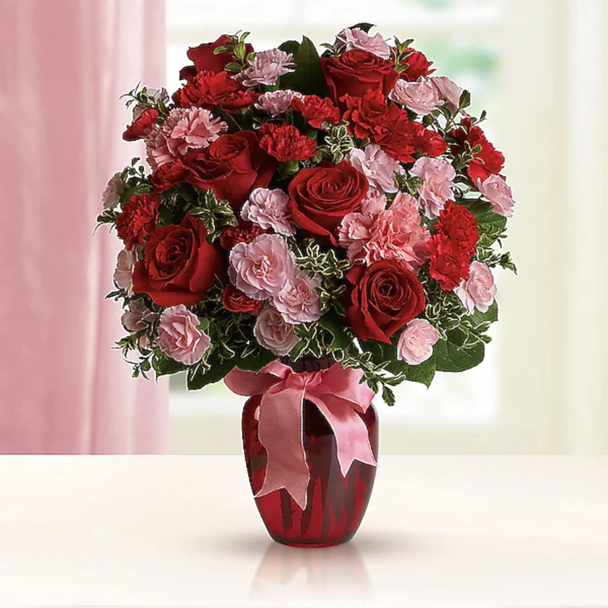 Red roses and pink carnations arranged in a red vase with a pink ribbon