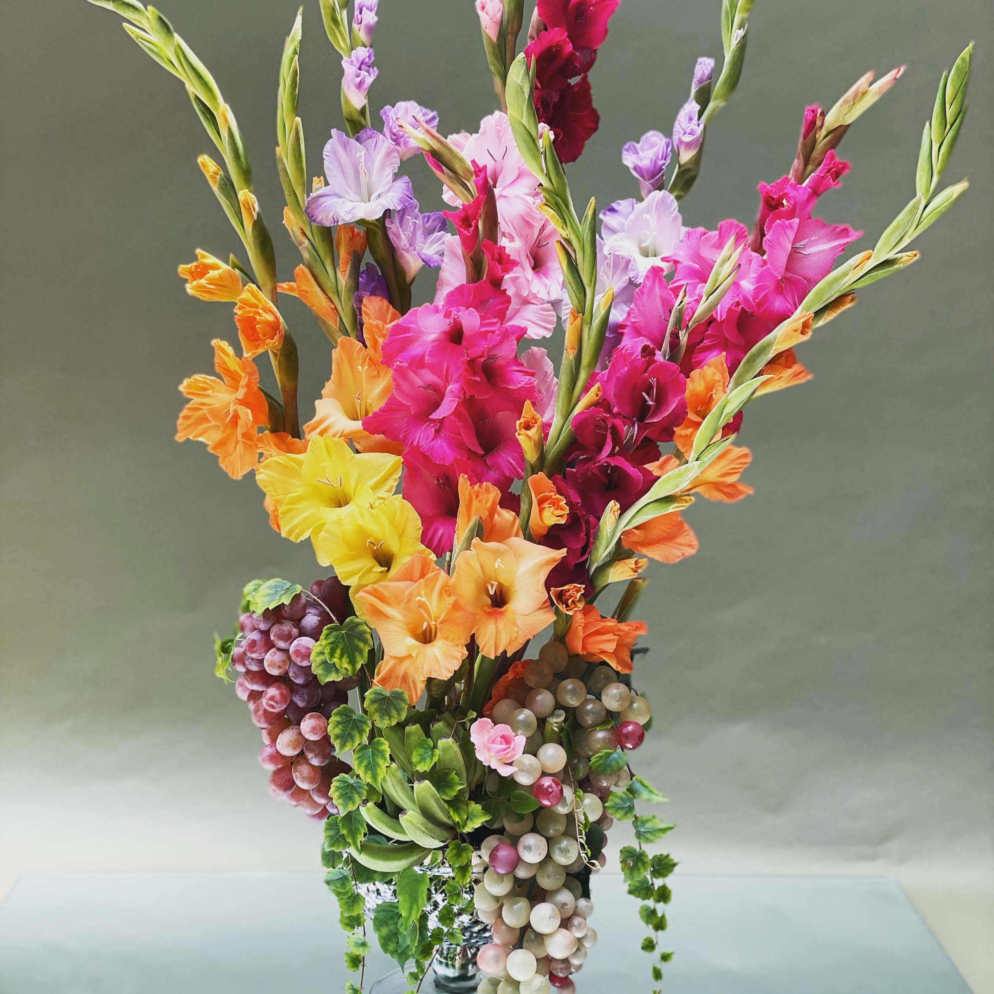 Tall bouquet of colorful gladiolus in a glass vase with hanging grape clusters