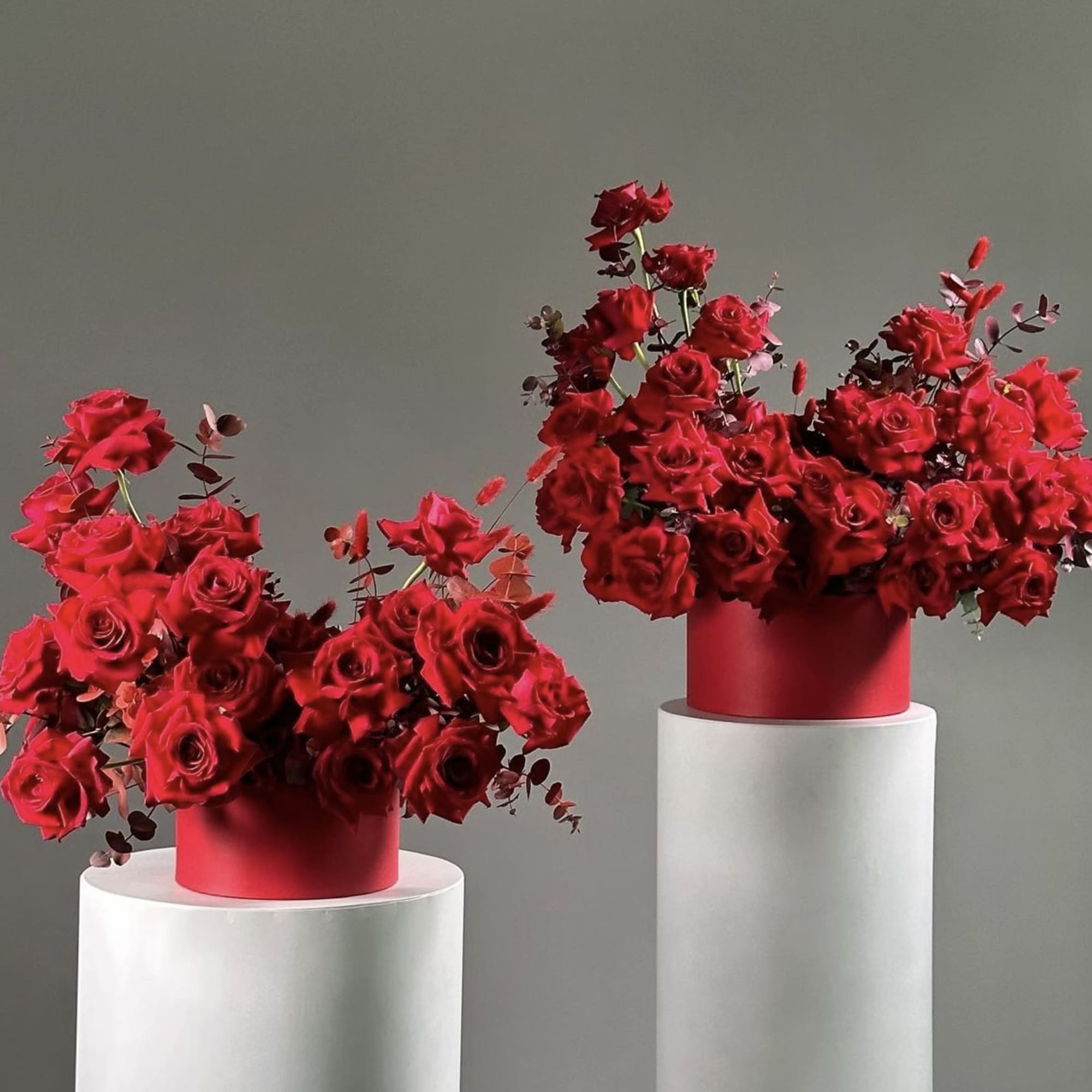 Two red rose arrangements in round red boxes on white pedestals