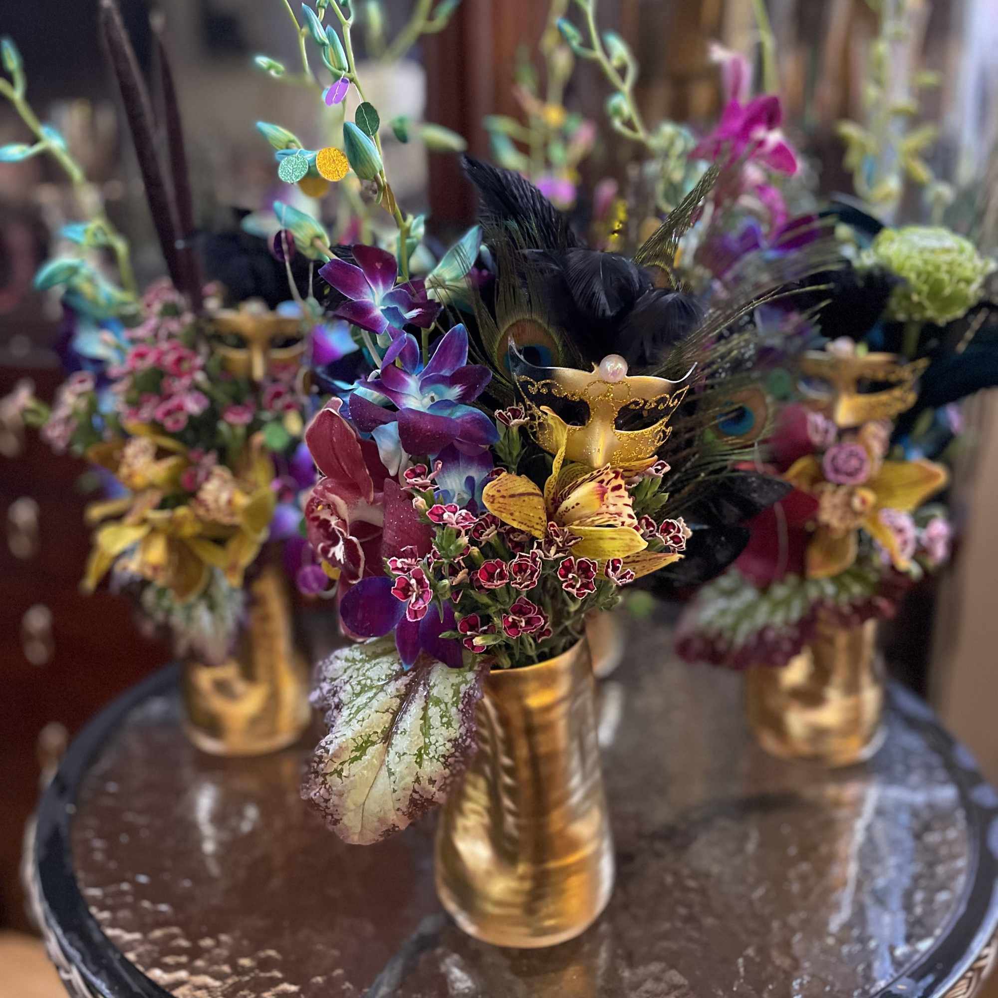 Orchid arrangement in a gold vase with black feathers and decorative masks