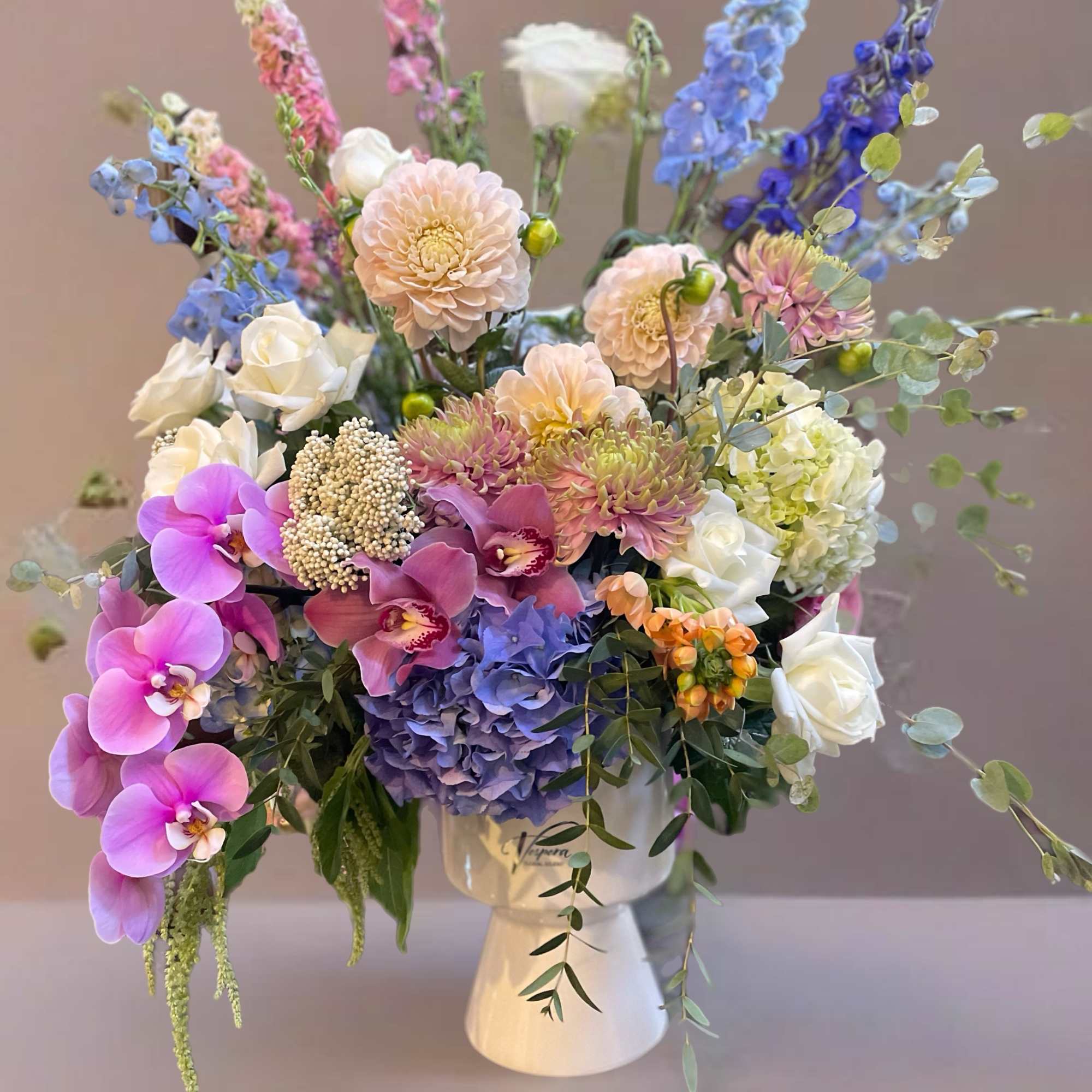 Mixed pastel bouquet in a white vase with orchids, roses, hydrangea, and tall blue blooms