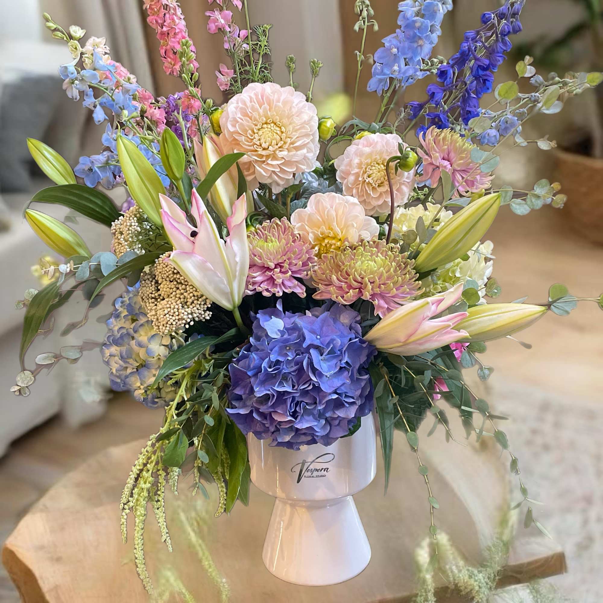 Pastel floral arrangement in a white pedestal vase with lilies, dahlias, hydrangea, and delphinium
