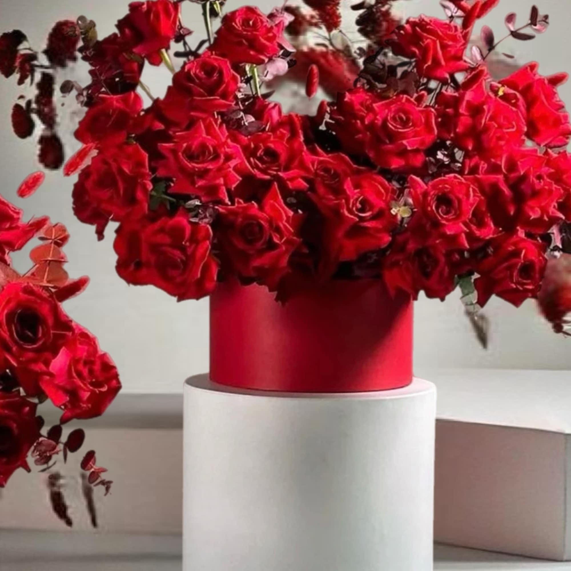 Red rose arrangement in a red cylindrical vase on a white pedestal, photo 2 of 2