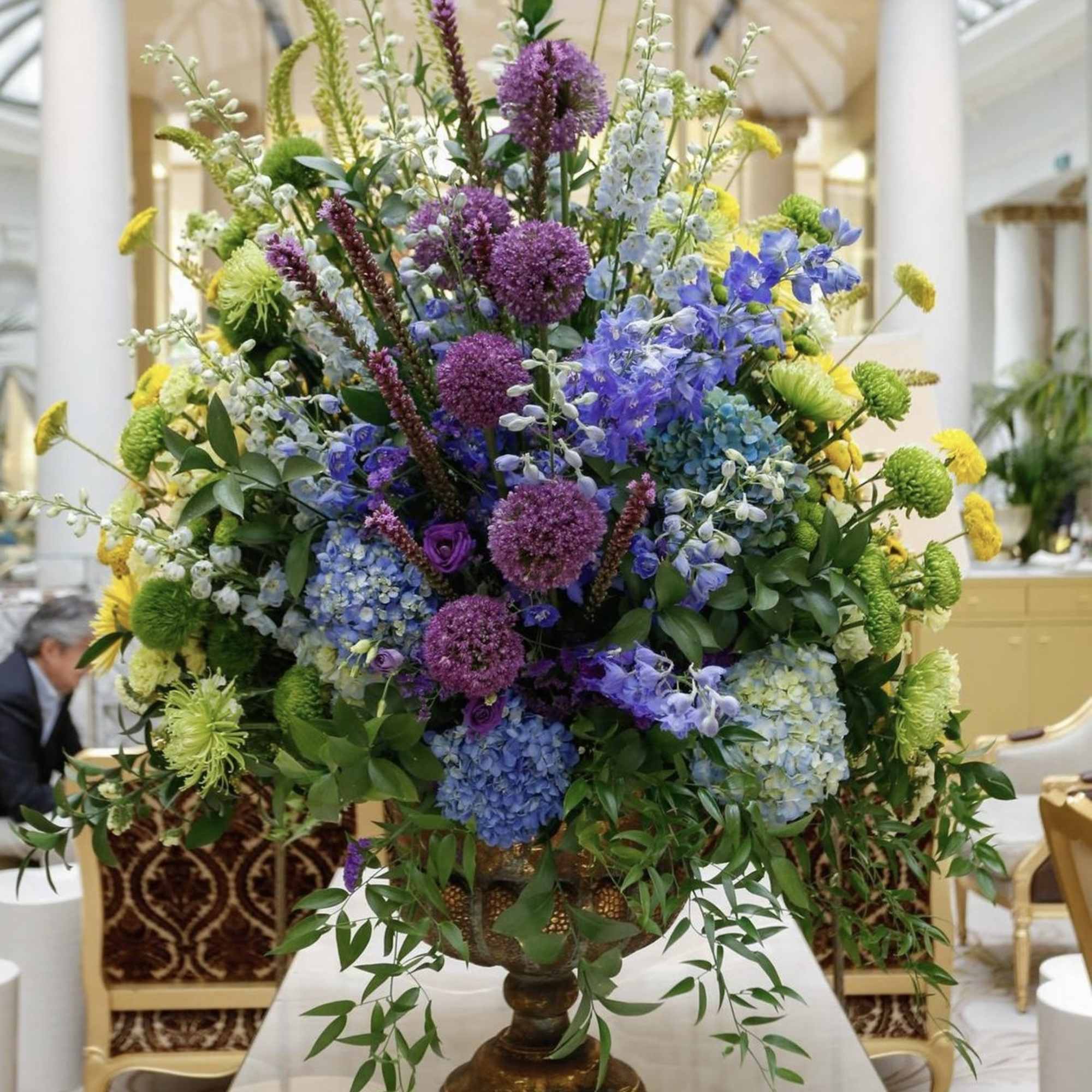 Large mixed floral centerpiece in a pedestal vase with purple, blue, yellow, and green blooms