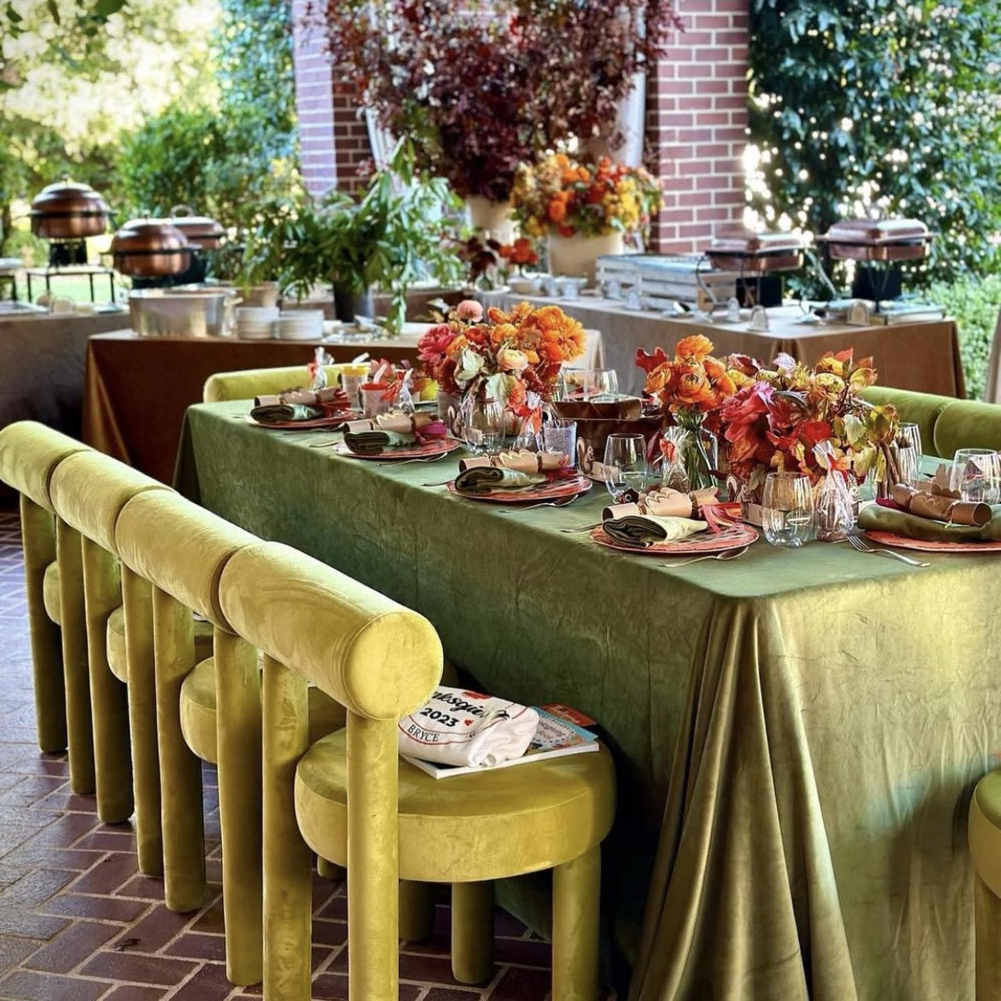 Outdoor banquet table with orange floral centerpieces and green velvet chairs