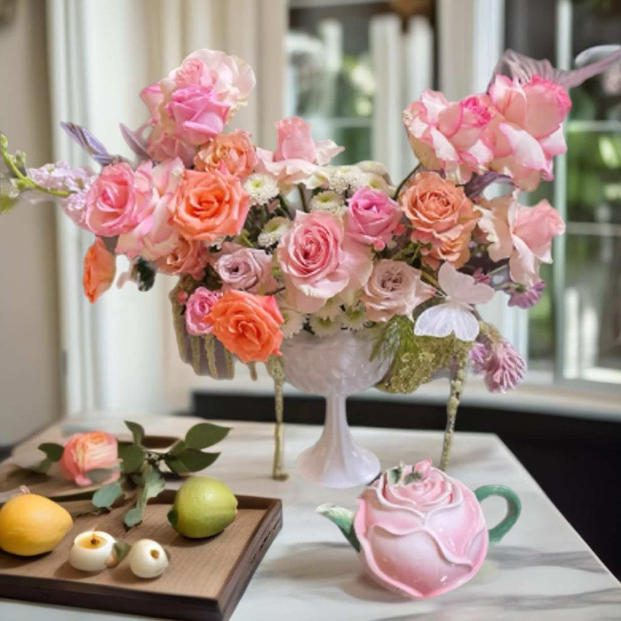 Pink and peach rose arrangement in a white pedestal vase with a rose-shaped teapot