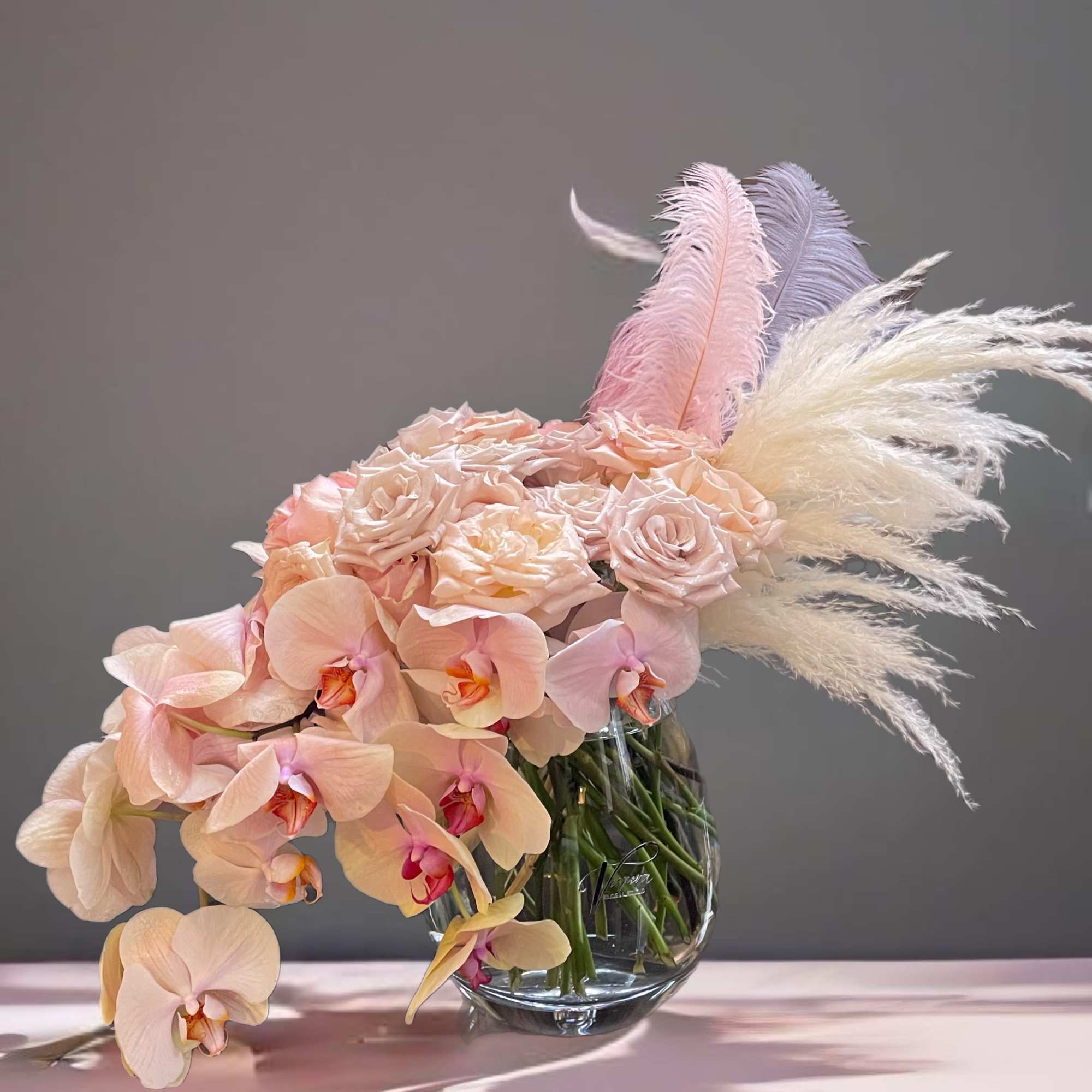 Blush orchids and pale roses in a clear glass vase with pastel feathers