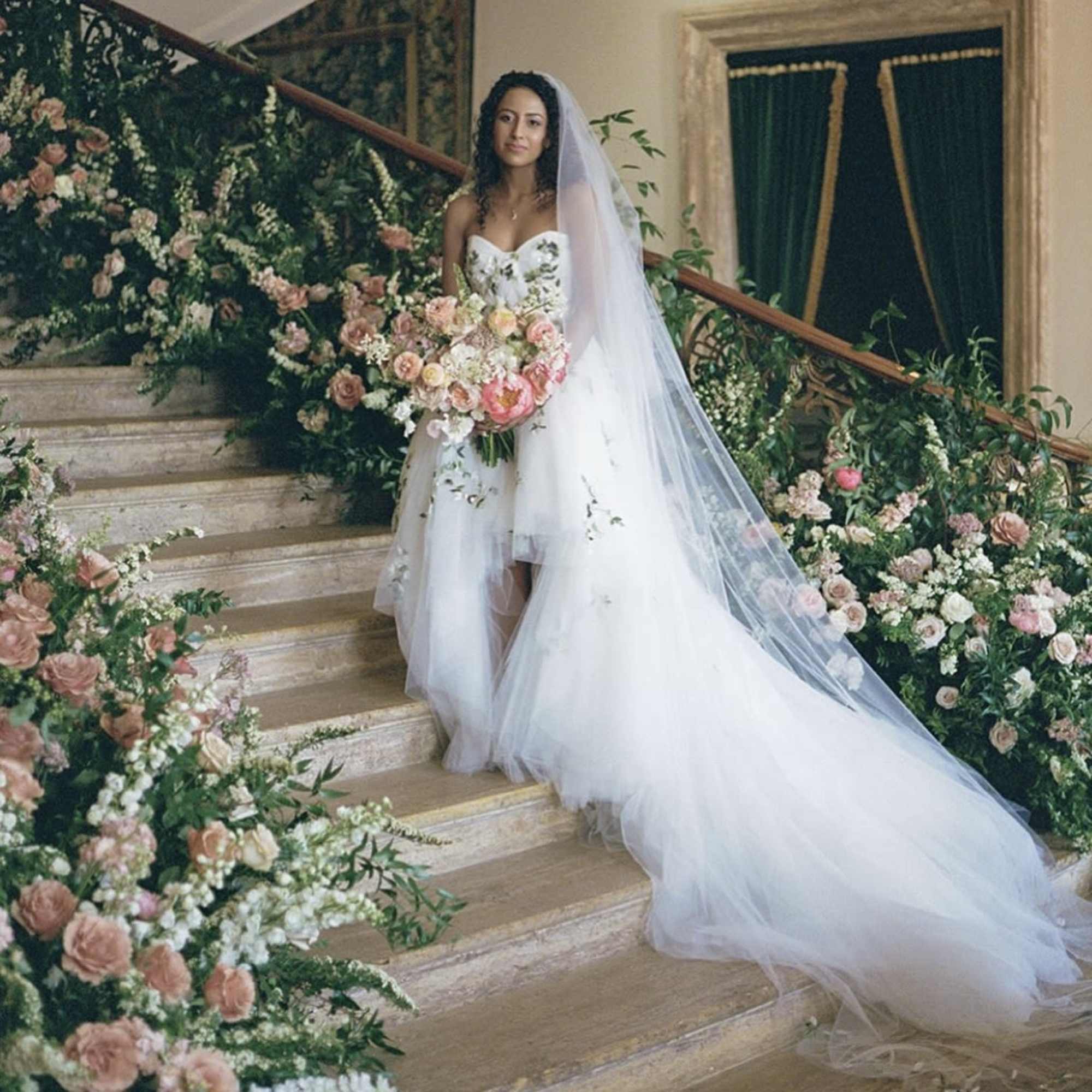 Bride in a white gown holding a pastel bouquet on a flower-lined staircase