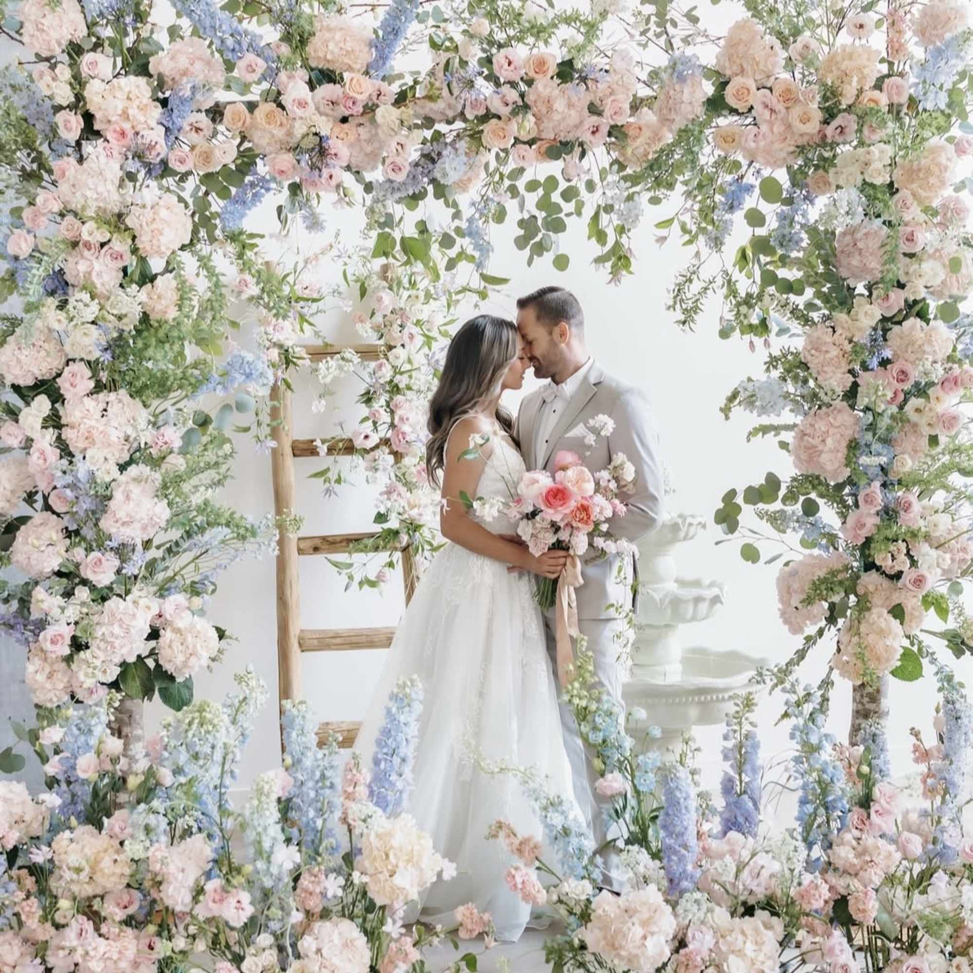 Bride and groom under a pastel floral arch with a bouquet