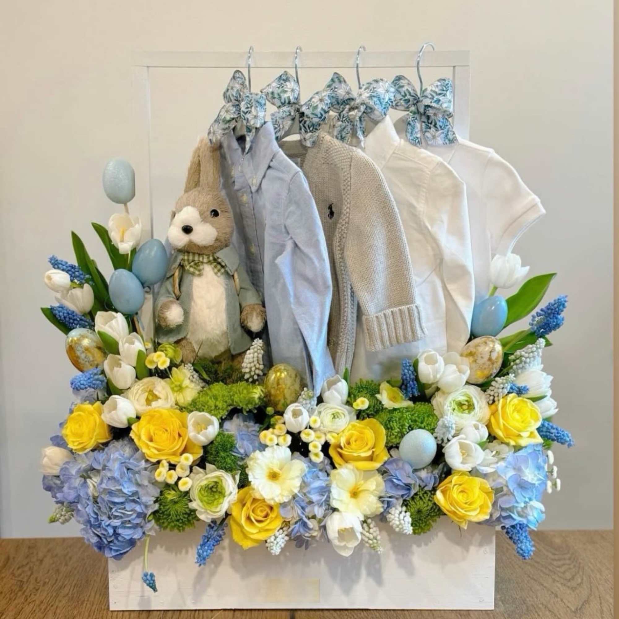 Baby clothes and a plush bunny surrounded by flowers in a white frame