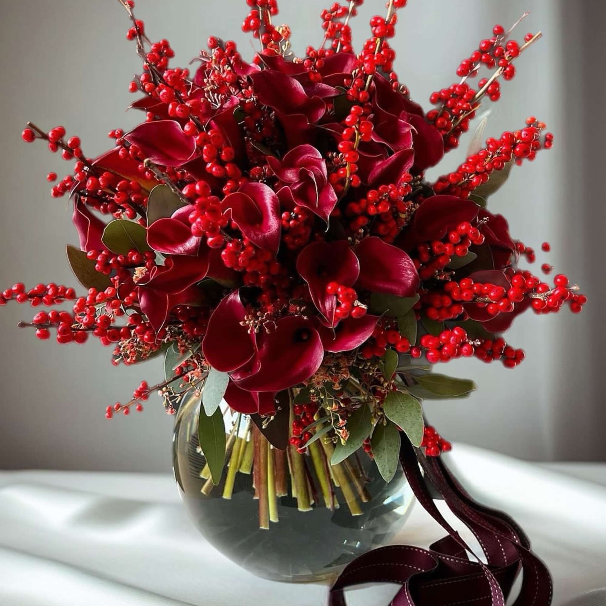 Deep red floral arrangement in a glass vase with berry branches and ribbon