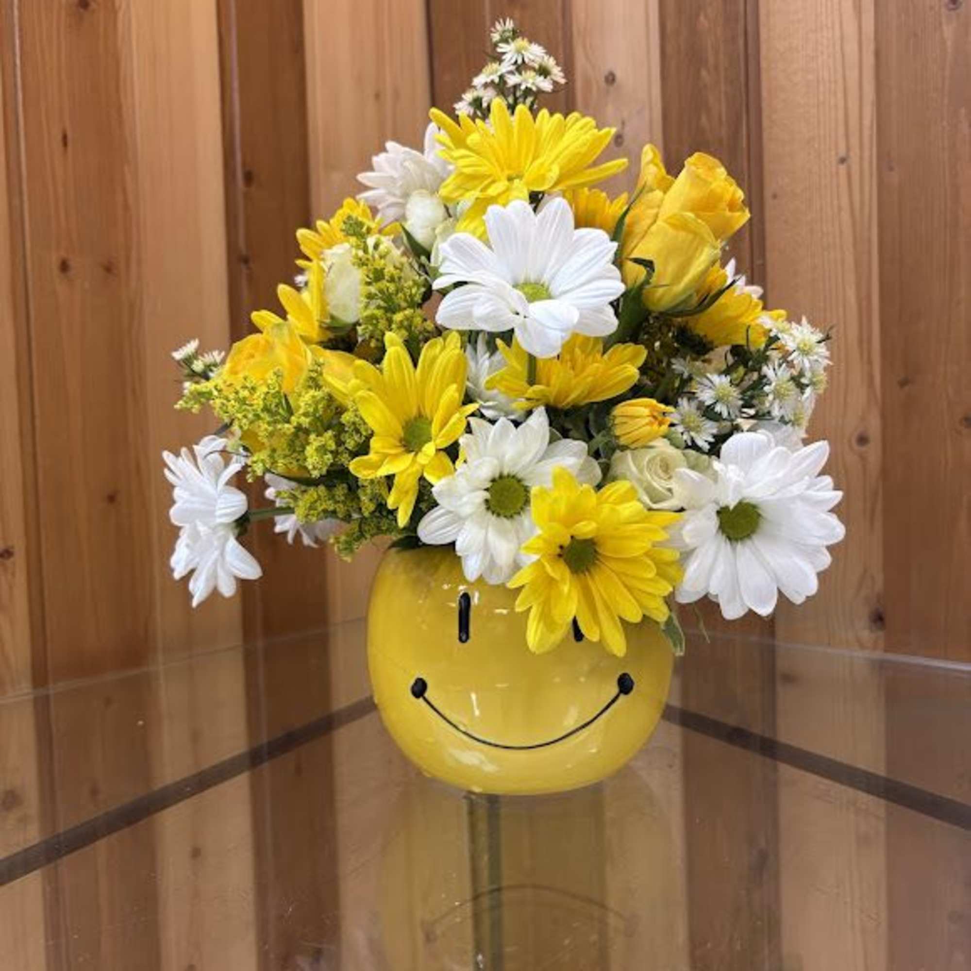 Yellow and white daisies with yellow roses in a yellow smiley-face mug on a glass table