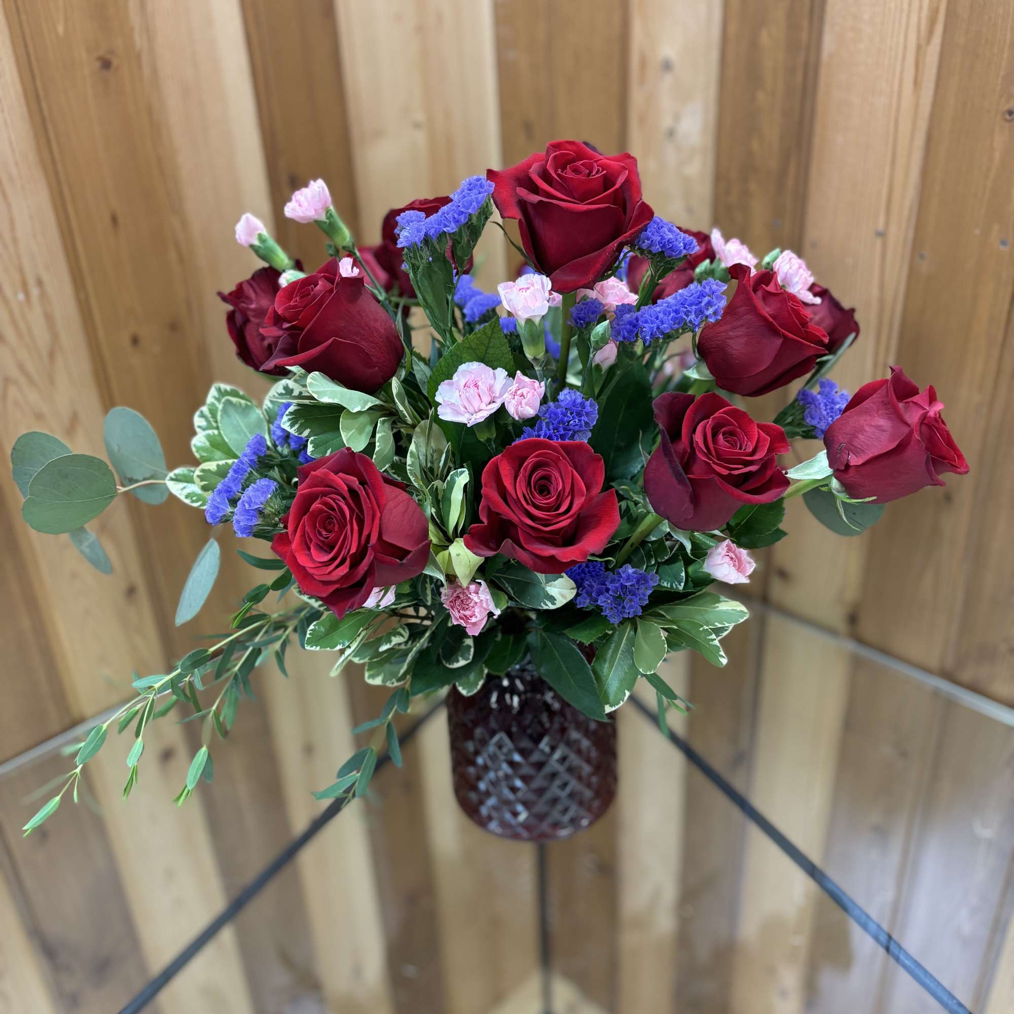 Arrangement of red roses, pink carnations, and purple filler flowers in a dark red glass vase.