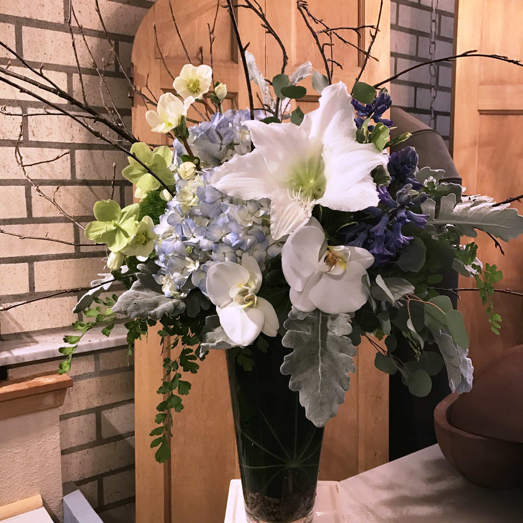 Tall floral arrangement with white orchids, hydrangea, and blue blooms in a dark vase