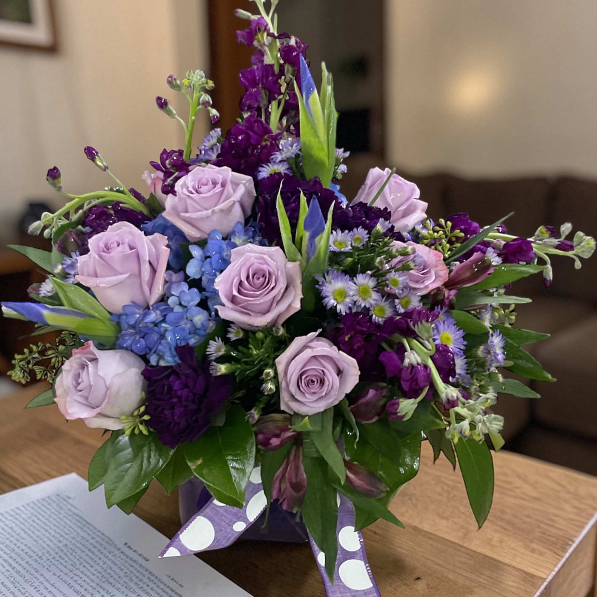 Lavender roses and deep purple blooms in a purple cube vase with a polka dot ribbon.