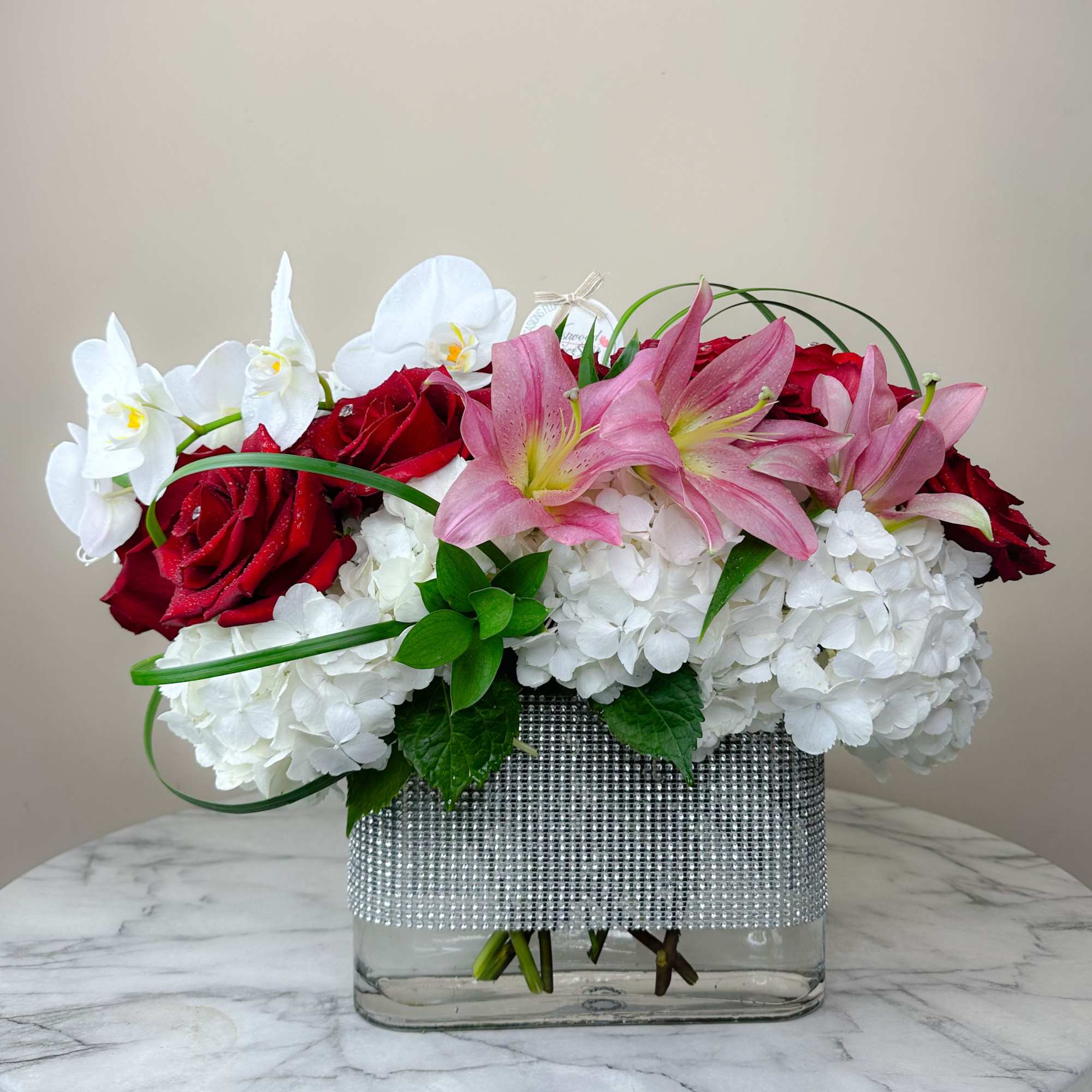 Pink lilies, red roses, and white orchids in a clear vase with rhinestone wrap