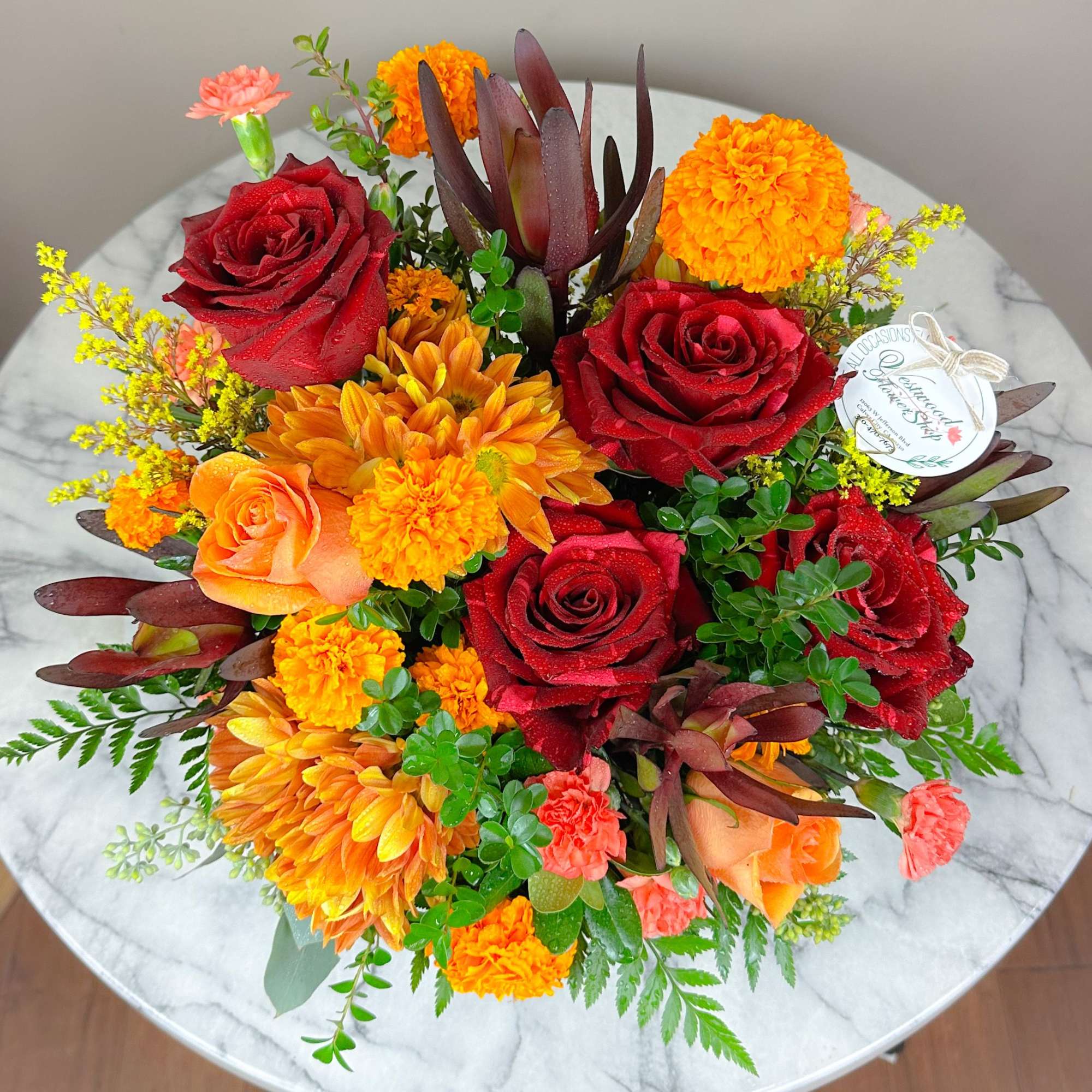 Round bouquet of red roses, orange blooms, and yellow accents on a marble table.