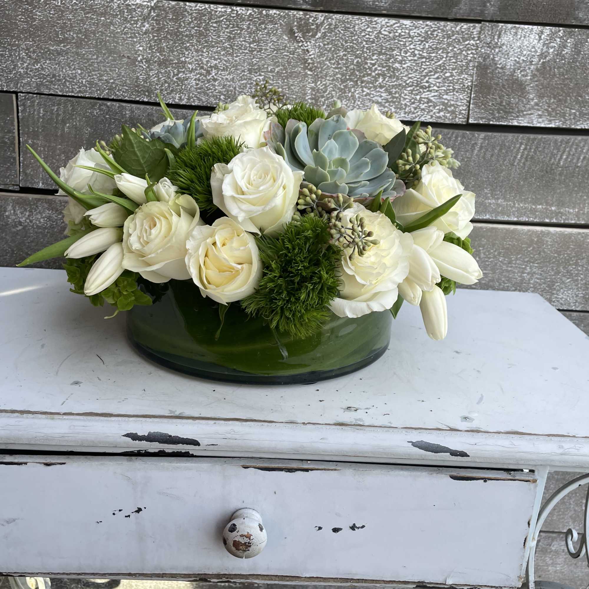 White roses and lilies arranged with succulents in a green glass bowl.