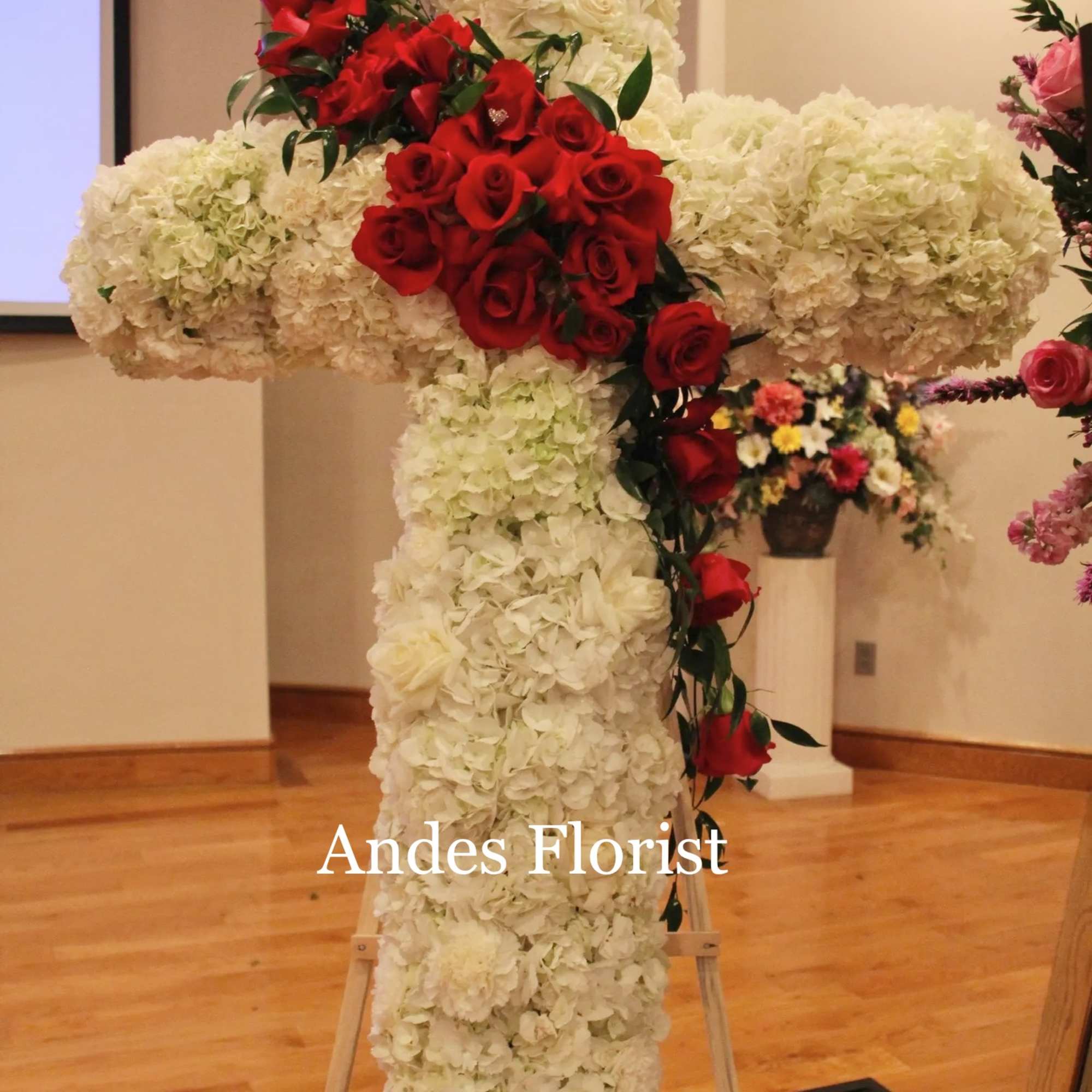 Large floral cross covered in white blooms with red roses draped across it