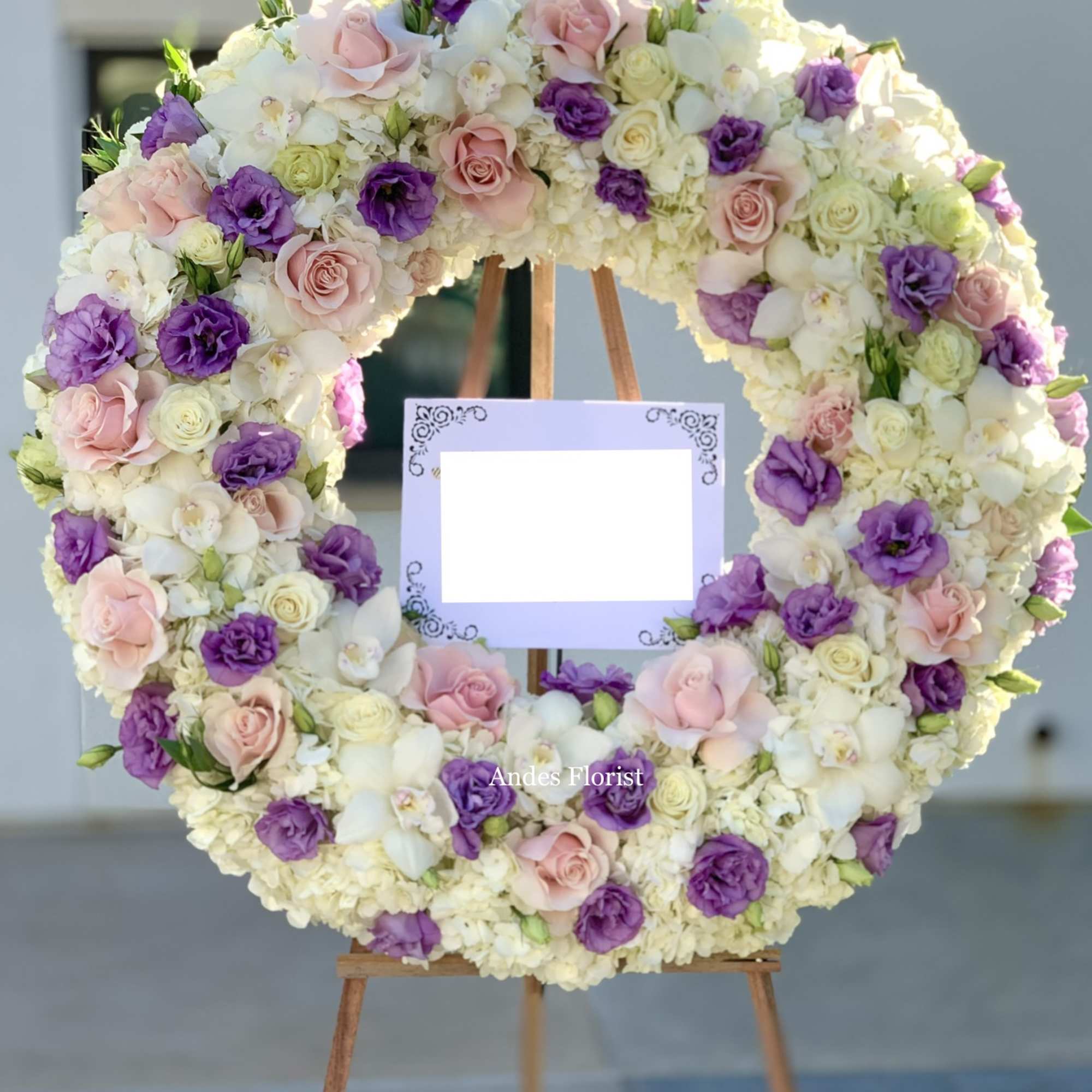 Large floral wreath of pink, purple, and white blooms on an easel with a blank sign