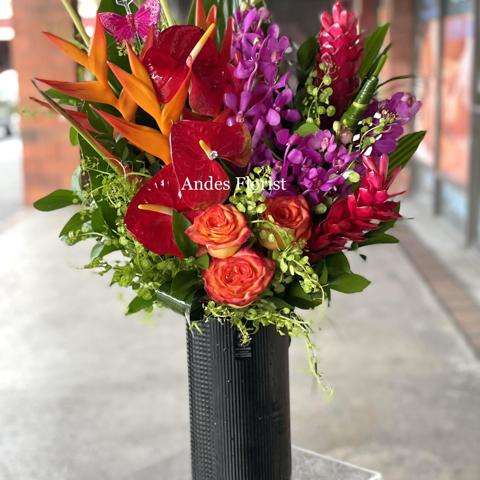Tropical bouquet with red anthuriums, orange birds of paradise, and purple orchids in a black vase.