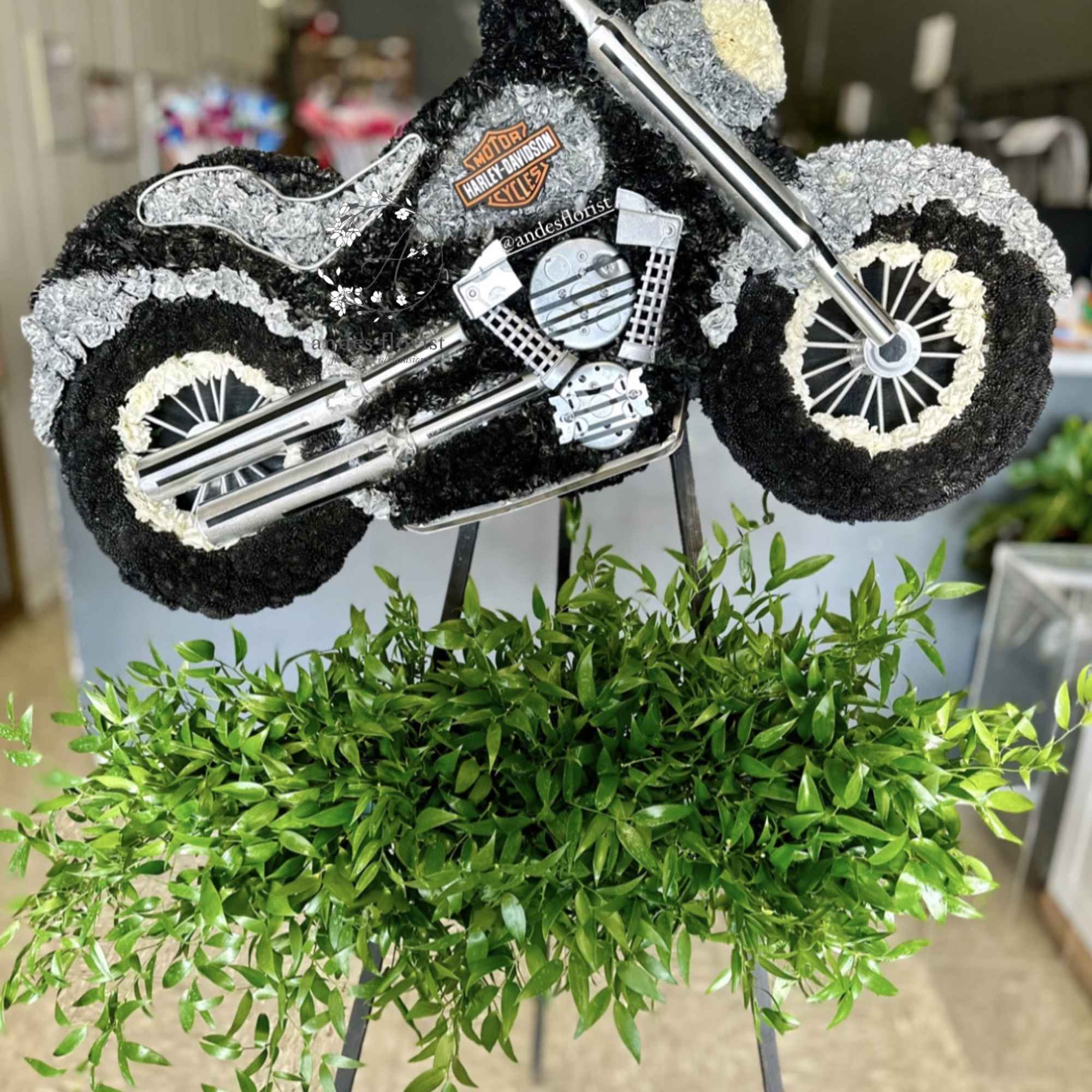 Motorcycle-shaped floral tribute with black, white, and gray flowers on an easel