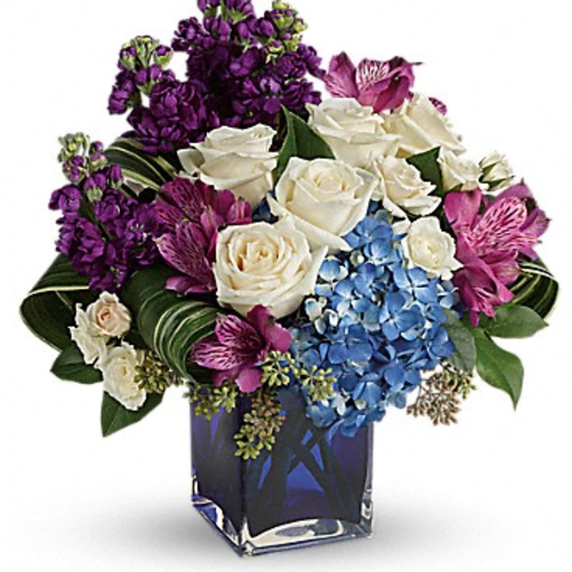 Bouquet of white roses, blue hydrangeas, and purple flowers in a square glass vase