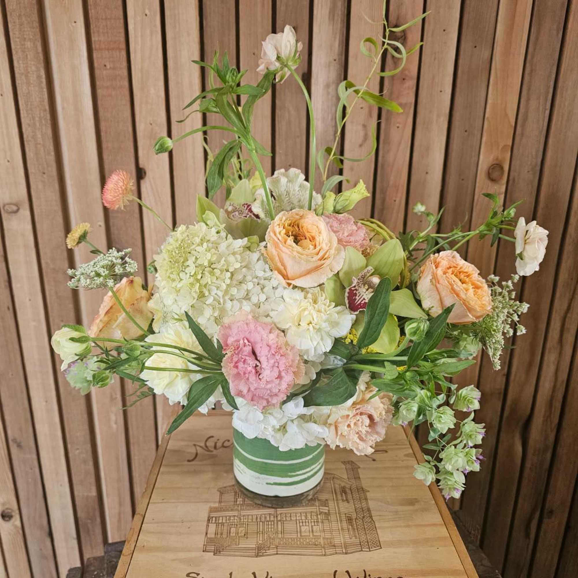 Loose pastel flower arrangement in a green-striped container on a wooden box