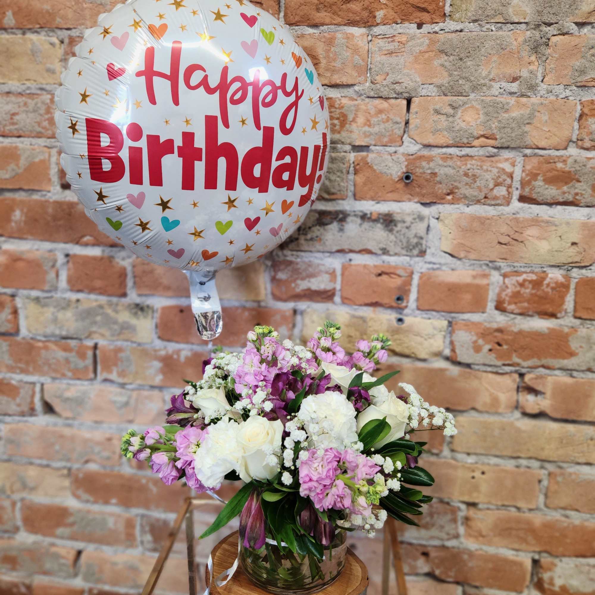Birthday balloon and small pink and white flower arrangement in a clear vase on a stand.