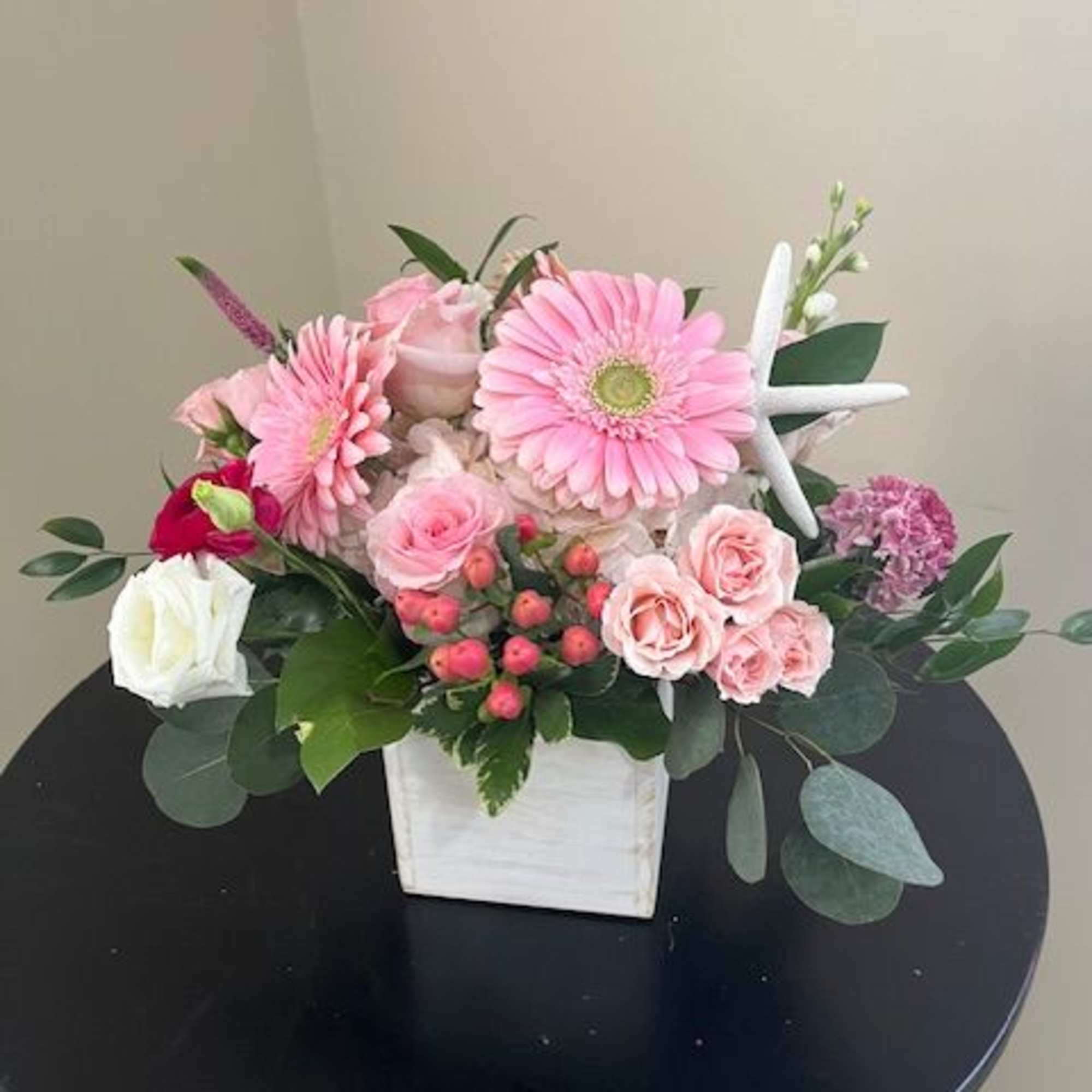 Pink gerbera daisies, roses, and spray roses in a white wooden cube with a decorative starfish