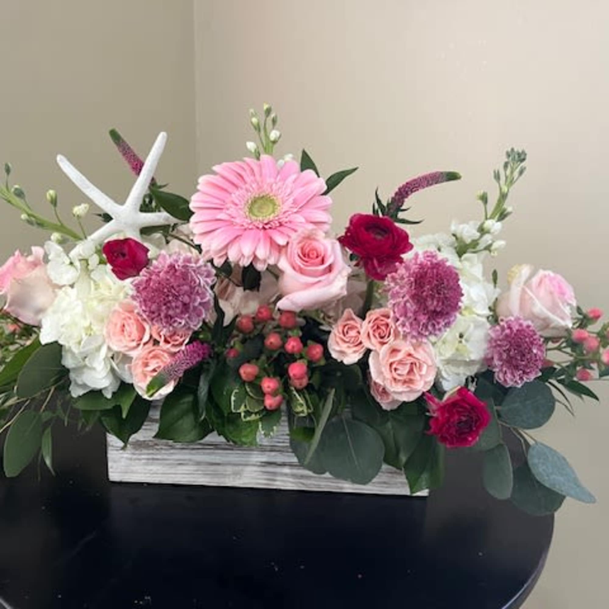 Low pink and white flower arrangement with gerbera, roses, and mums in a white wooden box with a decorative starfish.
