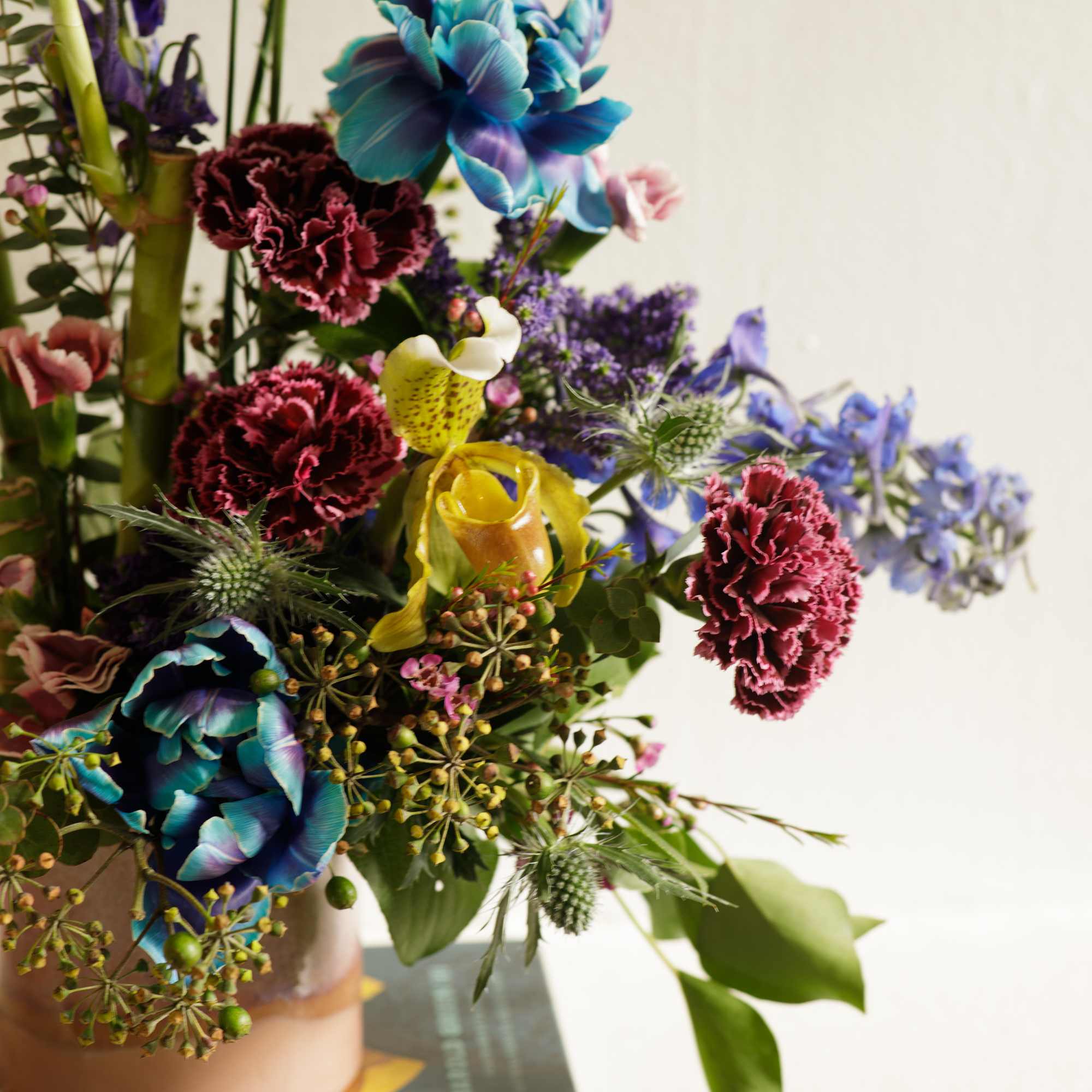 Colorful mixed arrangement with blue, burgundy, and yellow blooms in a blush vase set on a book.