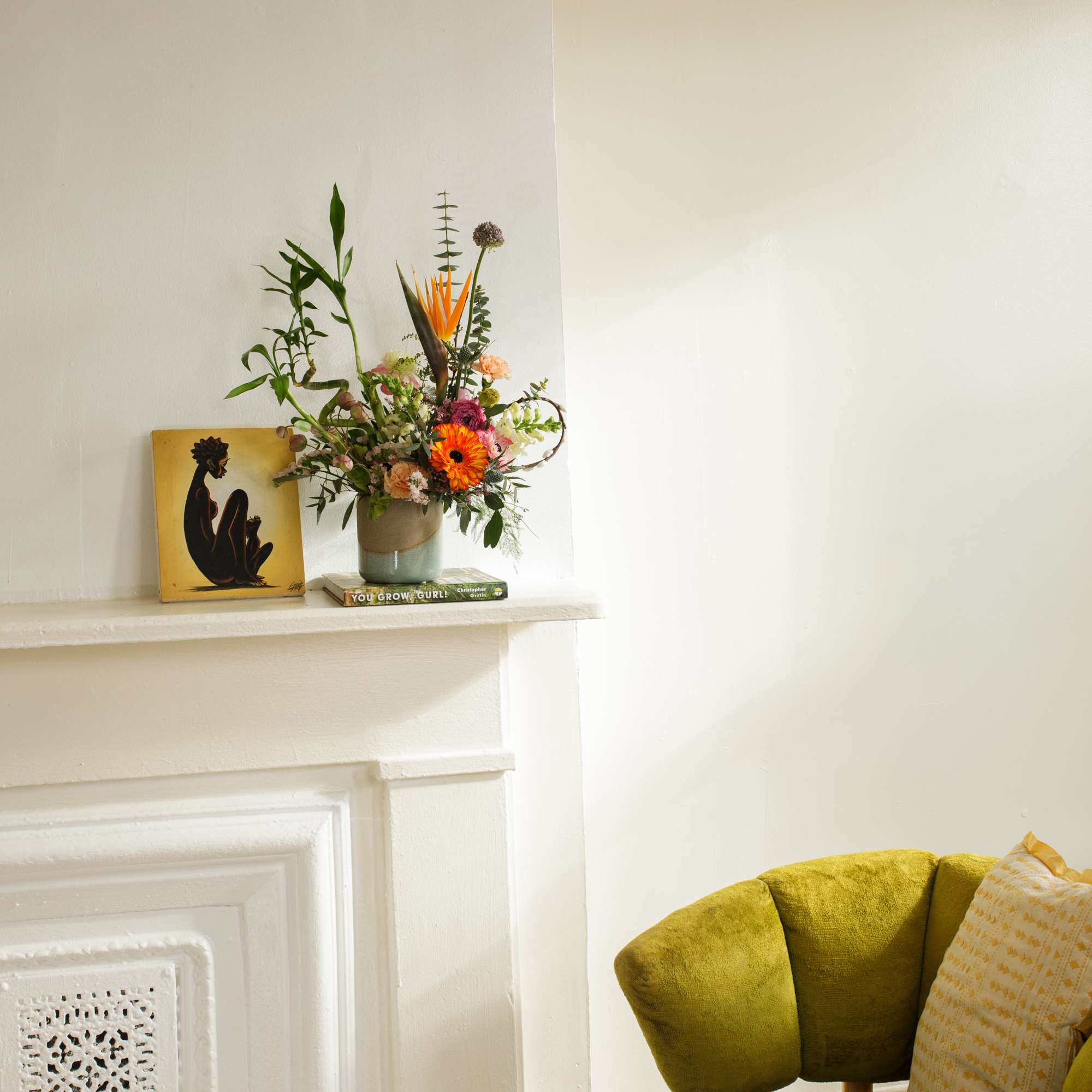 Colorful mixed flower arrangement in a ceramic pot on a white mantel beside artwork and a green chair.