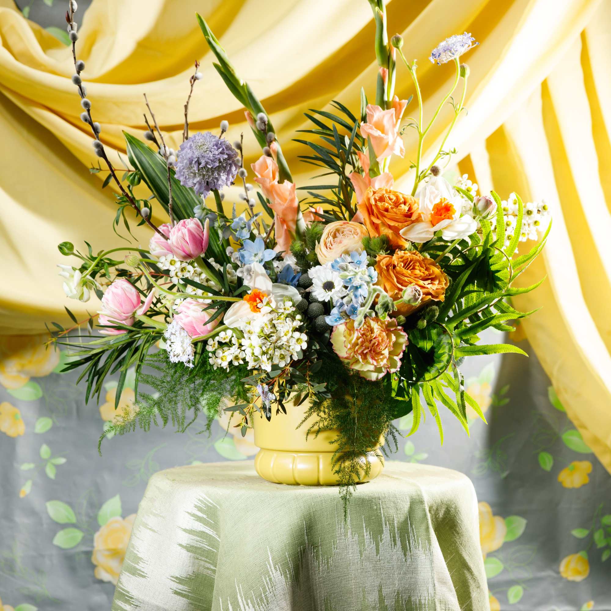 Tall mixed arrangement of peach, yellow, pink, and white flowers in a yellow vase on a draped table.
