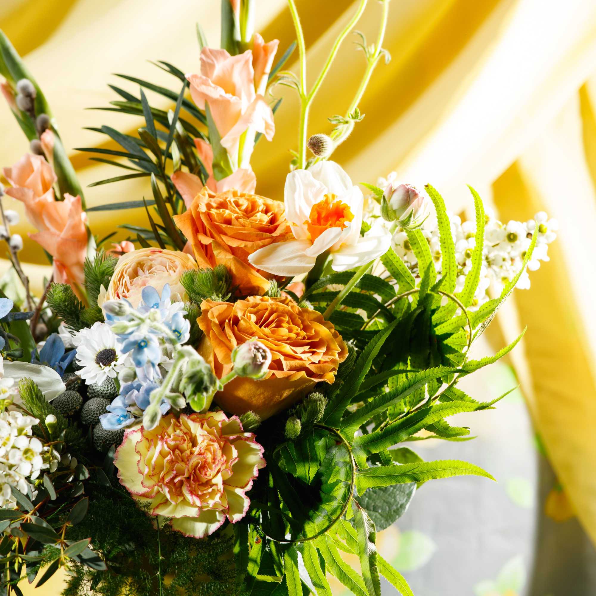 Bright mixed bouquet of orange roses, daffodils, and other blooms with ferns in a yellow vase