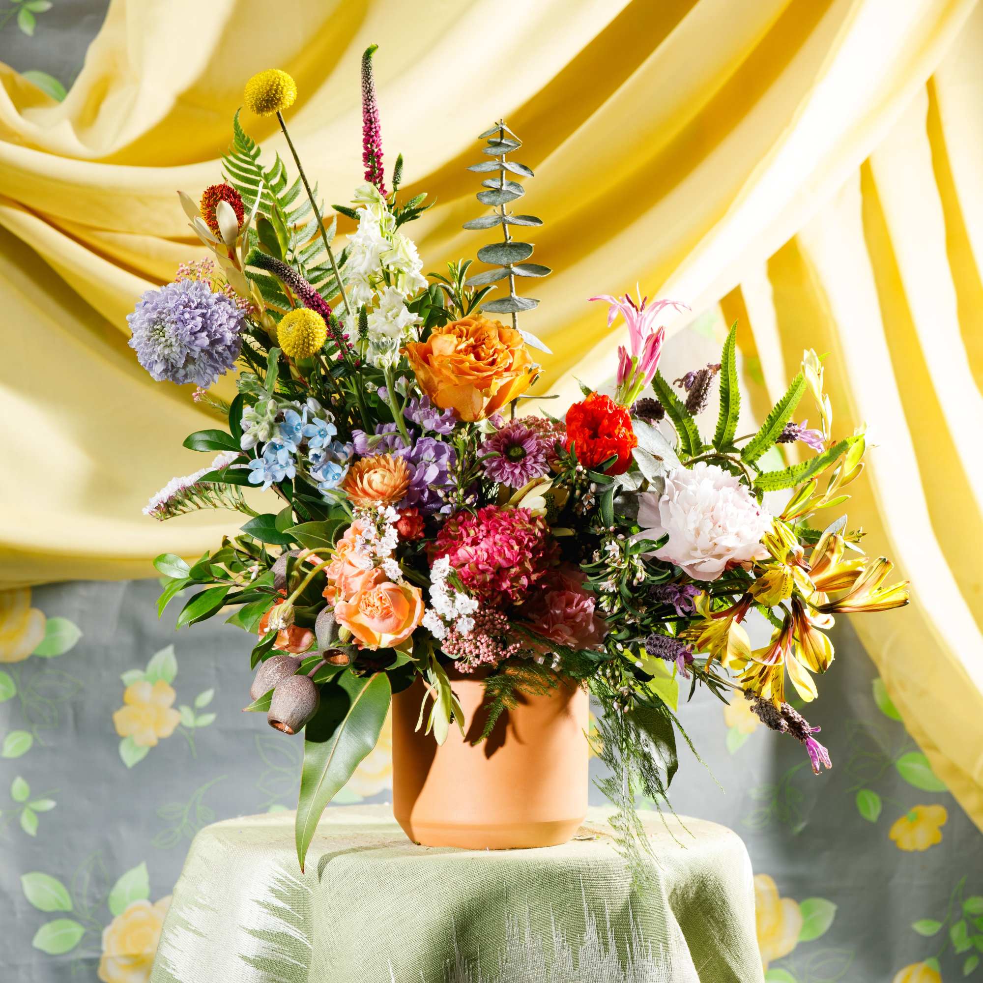 Colorful mixed flower arrangement in a terracotta pot on a draped table with yellow fabric backdrop.