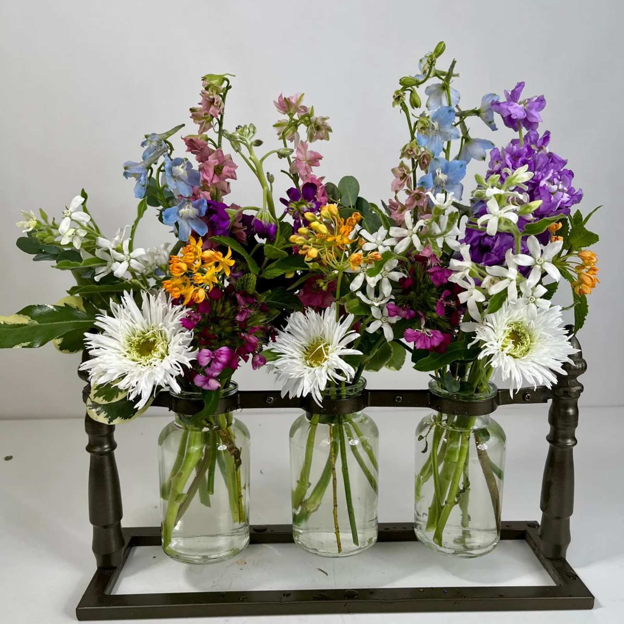 Three small glass jars hold mixed colorful flowers in a metal stand.