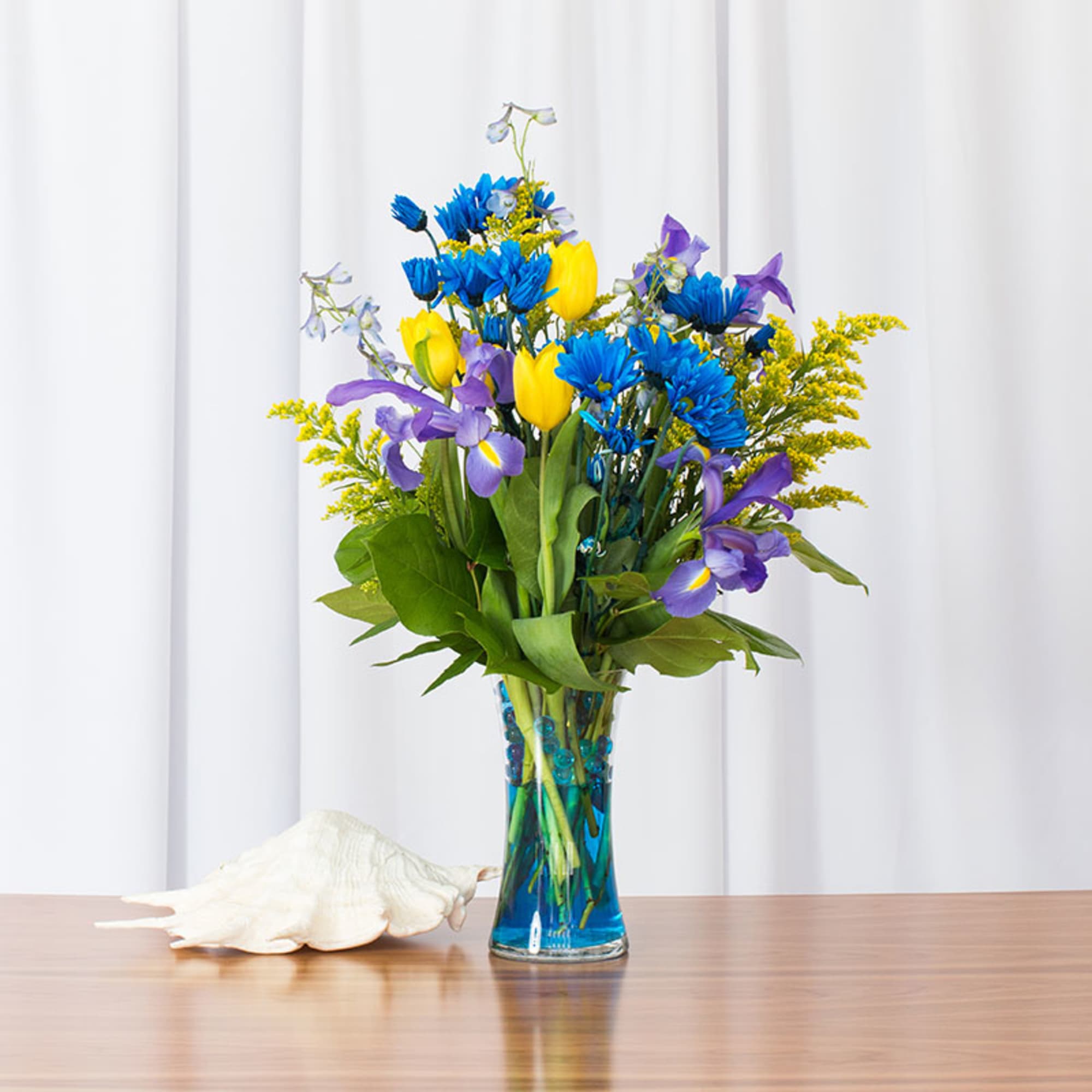 Tall vase of blue daisies, yellow tulips, and purple irises on a table with a white seashell.