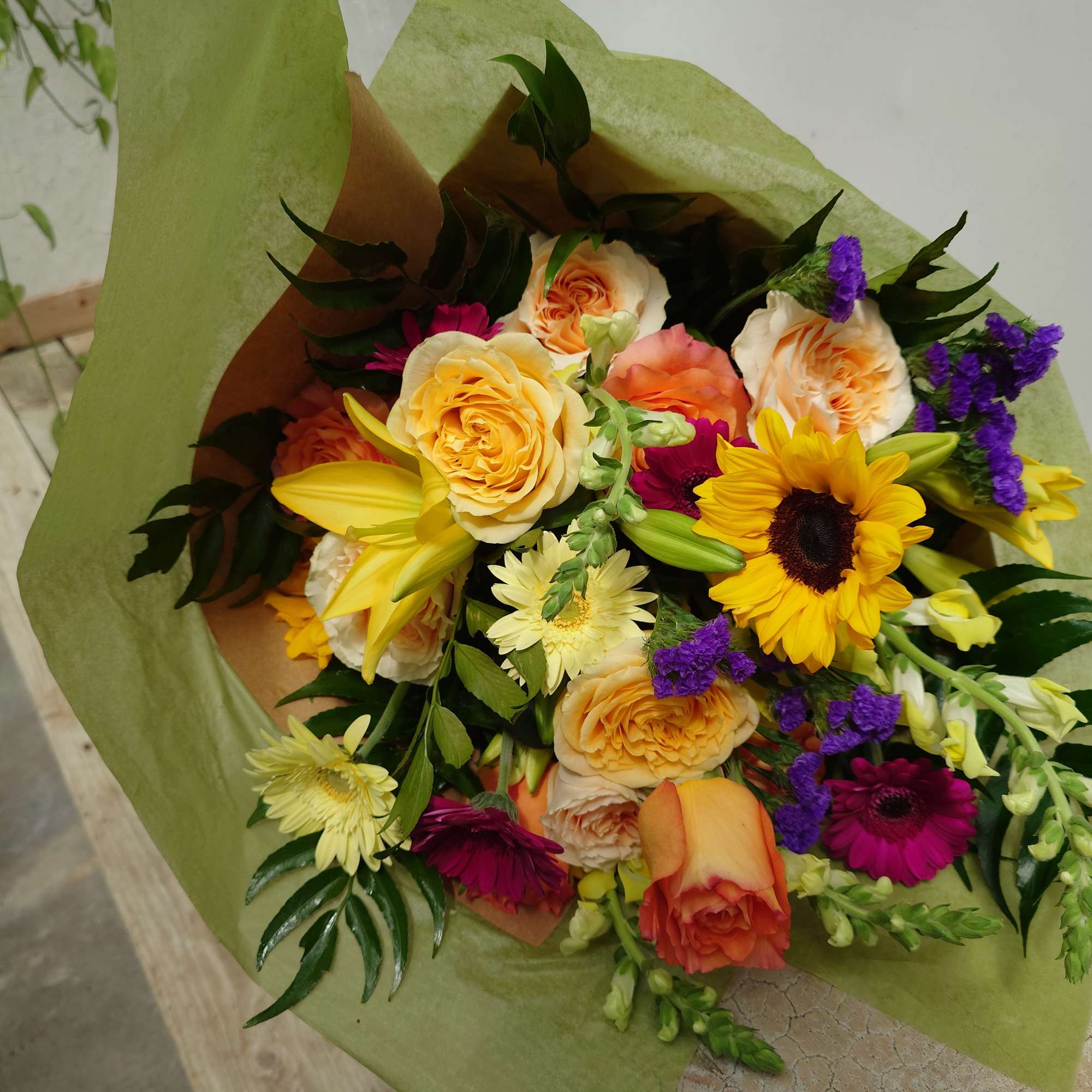Mixed bouquet of roses, sunflowers, lilies, and purple filler flowers wrapped in green paper