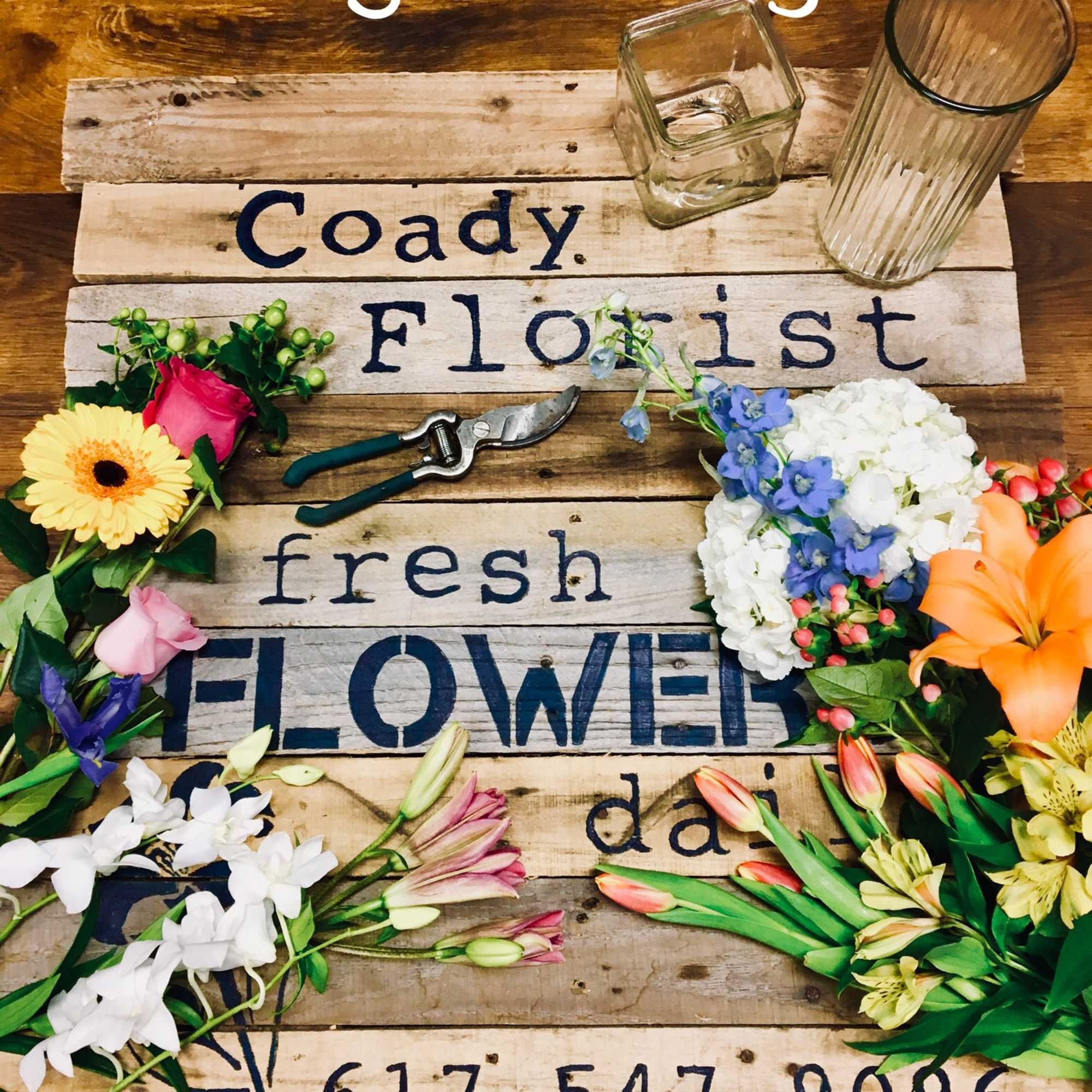 Assorted flowers arranged around a wooden florist sign with glass vases