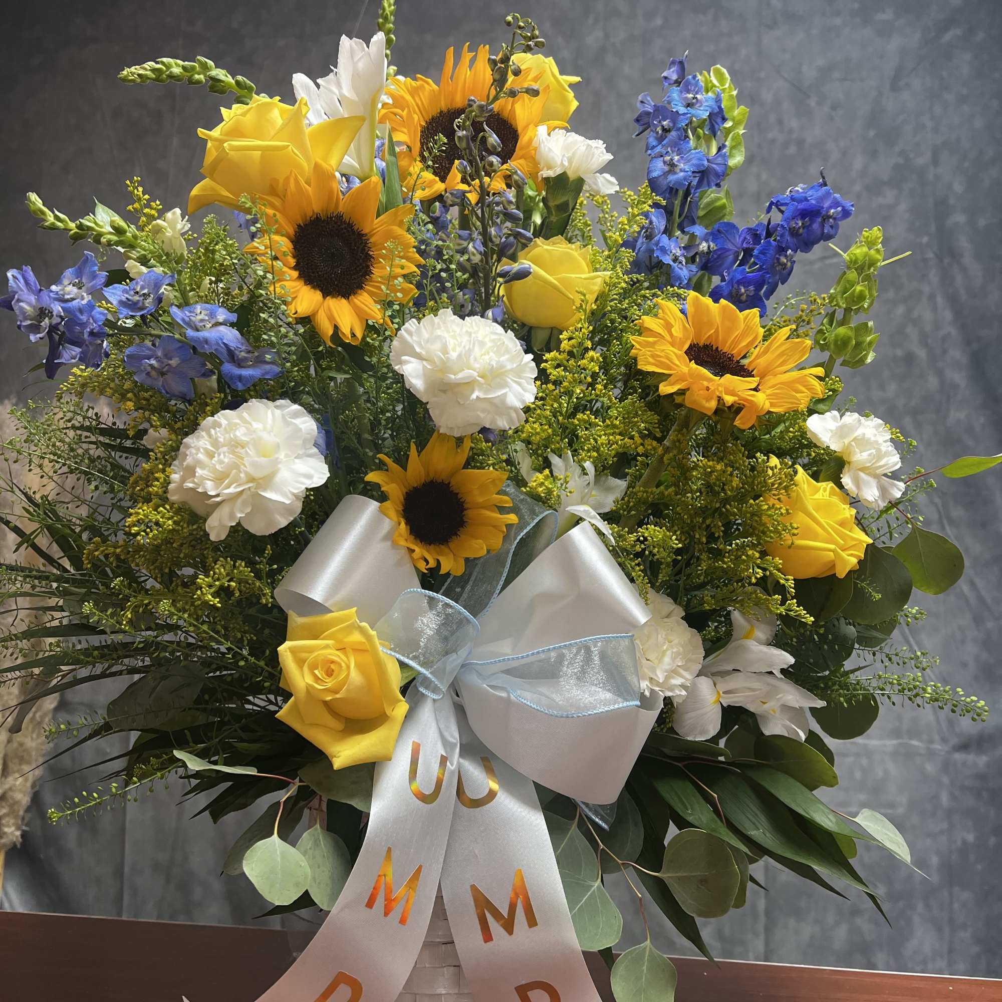 Basket arrangement of yellow roses, sunflowers, white carnations, and blue delphinium