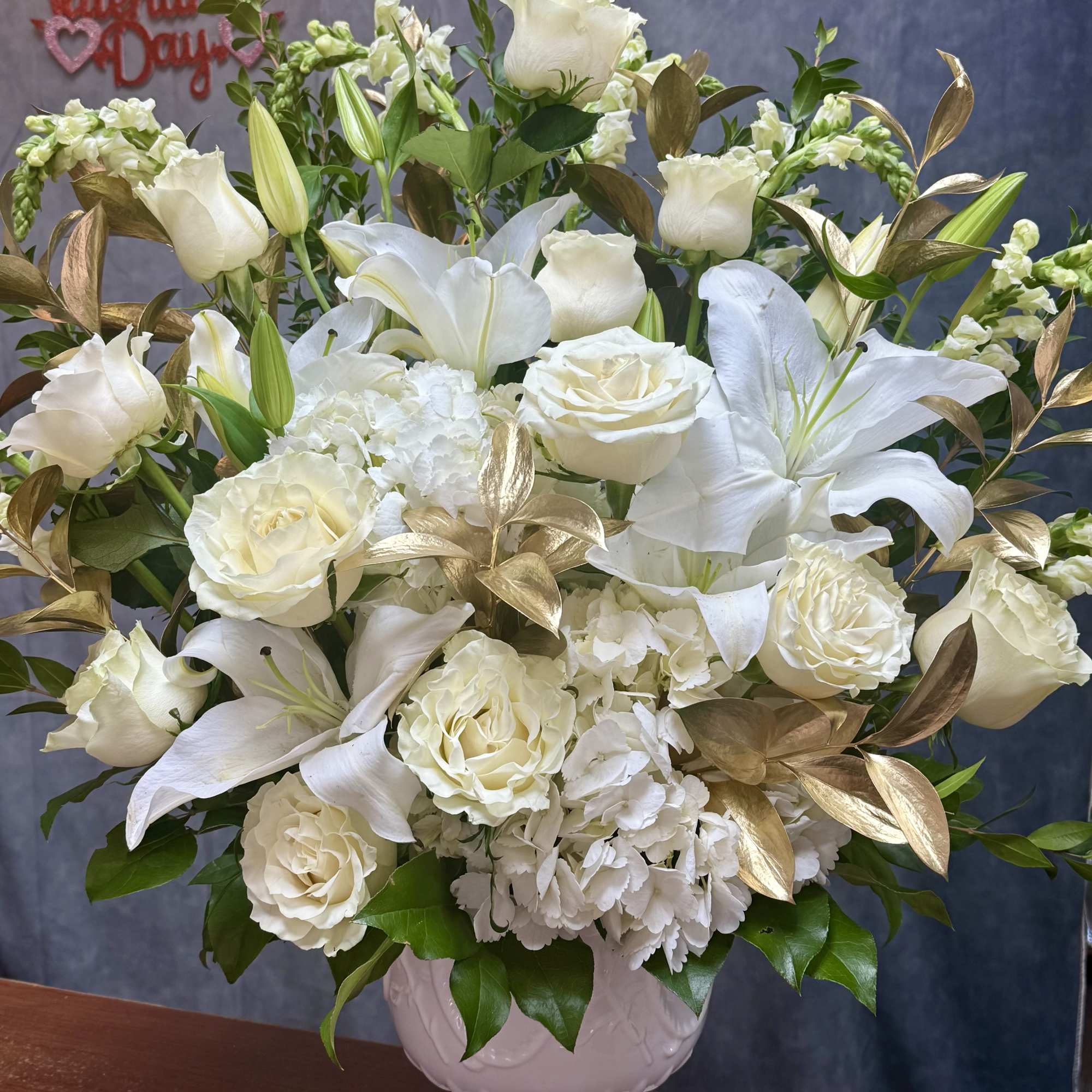 White roses and lilies arranged in a white vase with gold foliage accents.