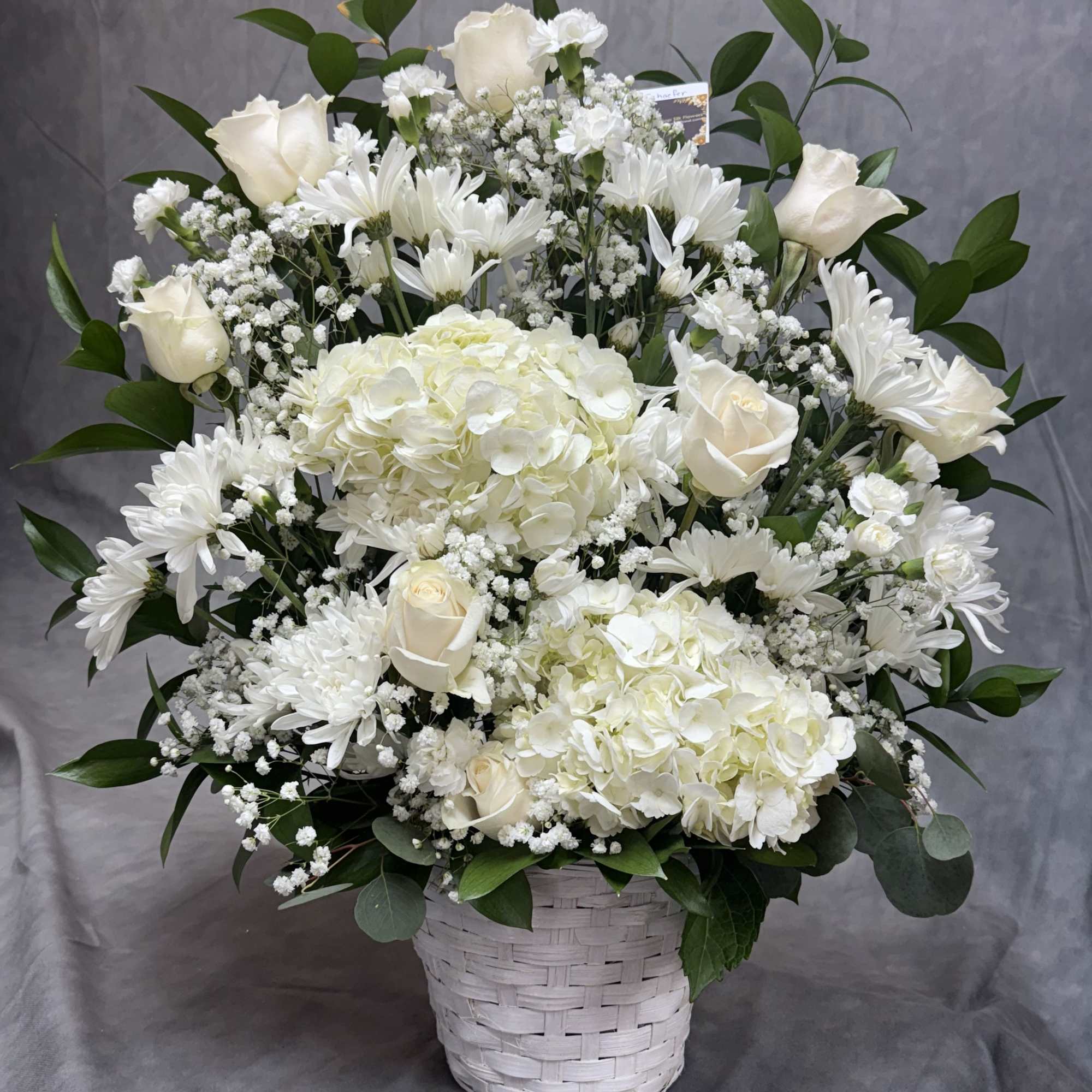 White floral arrangement in a woven basket with roses, hydrangeas, and daisies