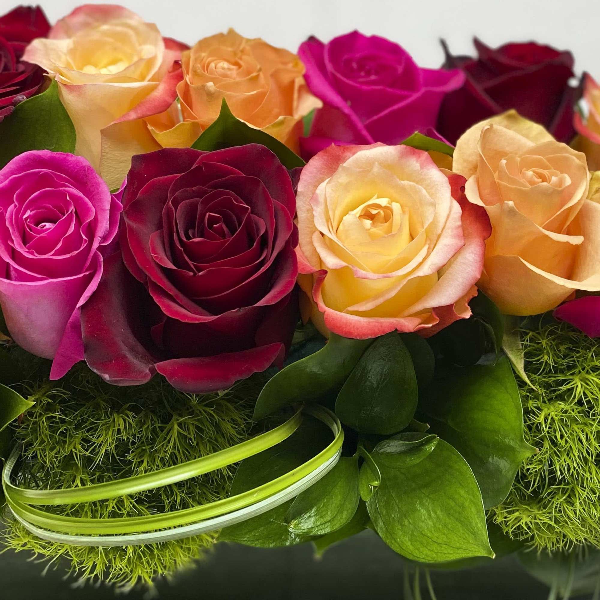 Low arrangement of hot pink, red, and peach roses set above green moss in a clear glass vase.