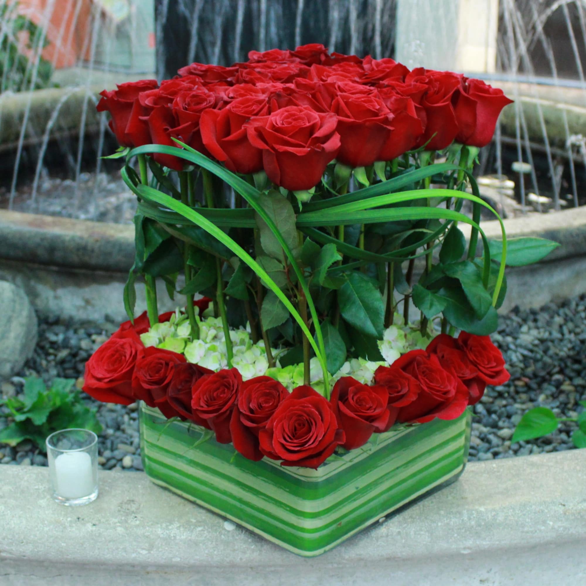 Red roses arranged in a striped green square vase with a candle beside it.