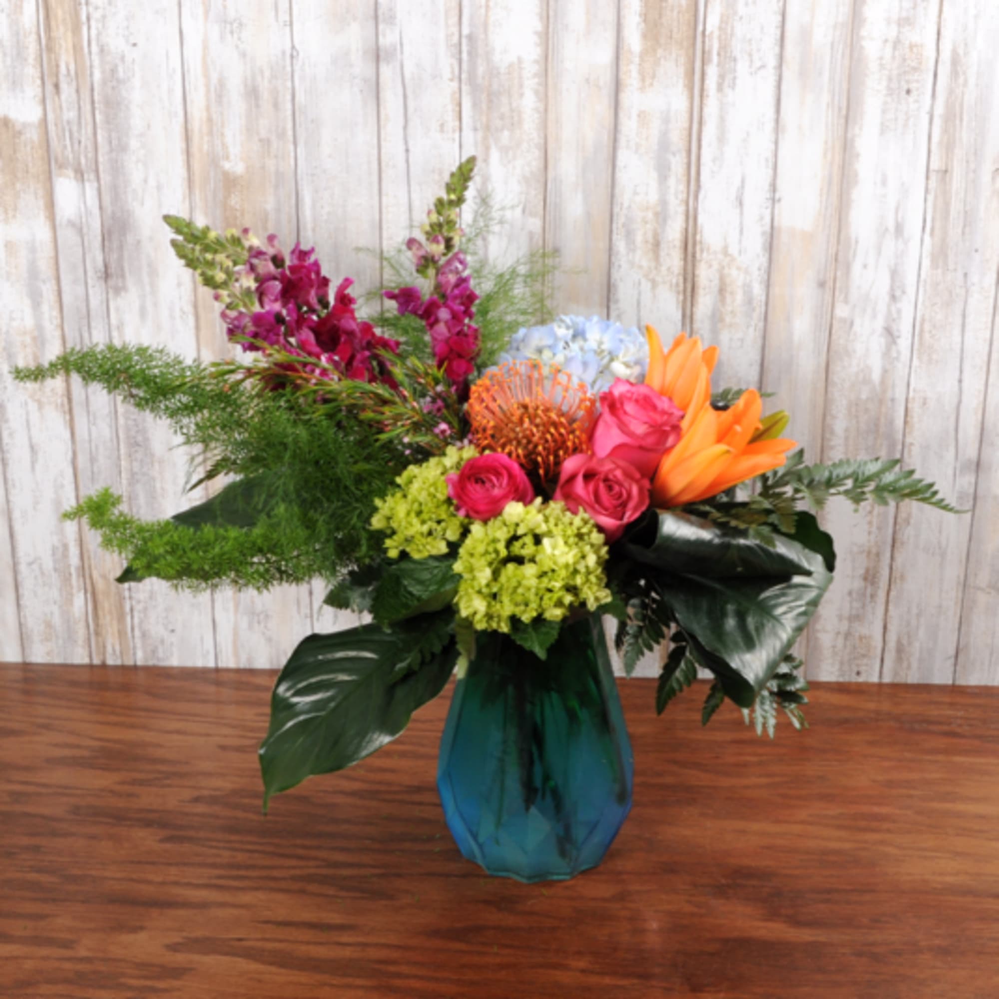 Colorful arrangement with pink roses, orange lilies, hydrangeas and protea in a blue glass vase