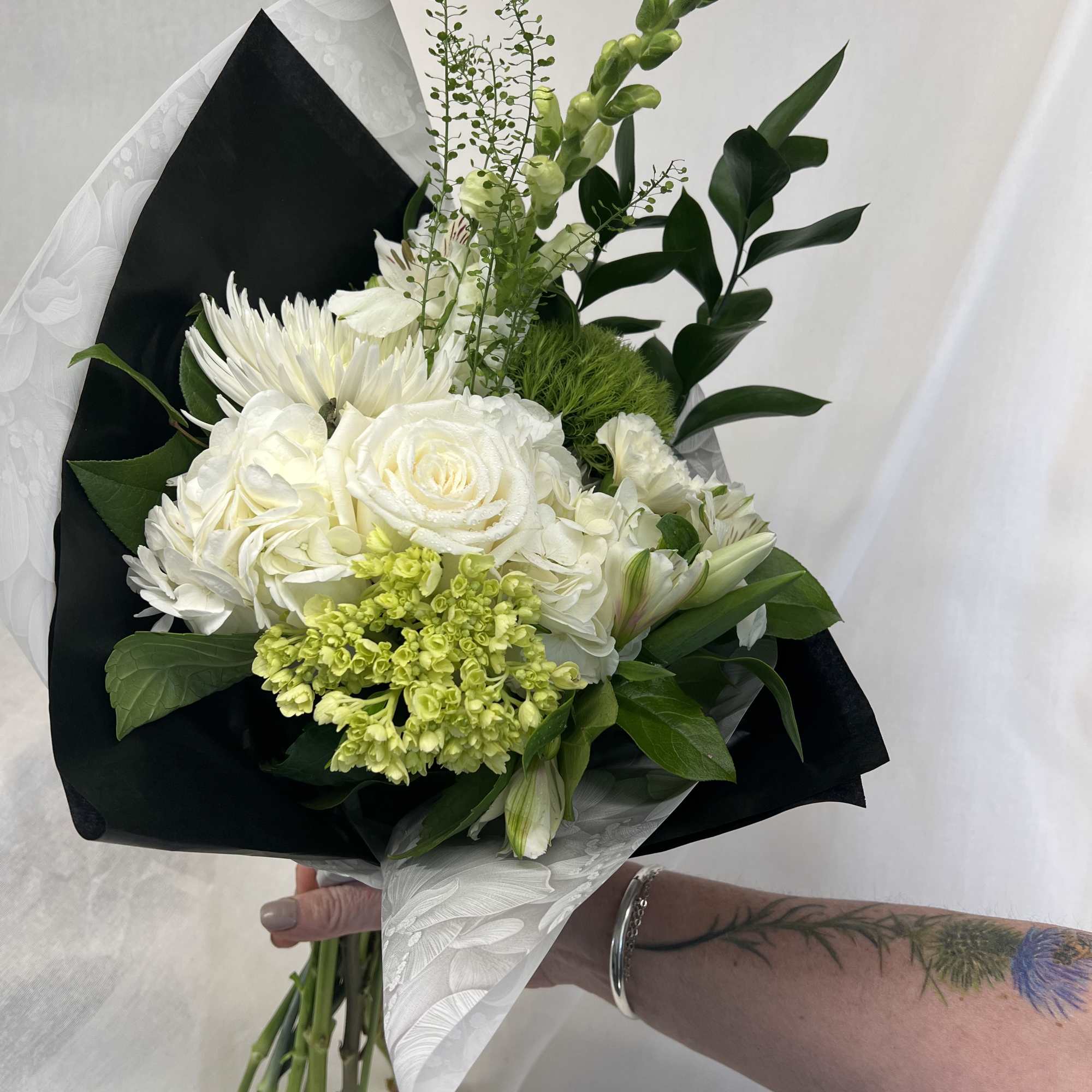 White bouquet with roses, chrysanthemums, and green accents wrapped in black paper