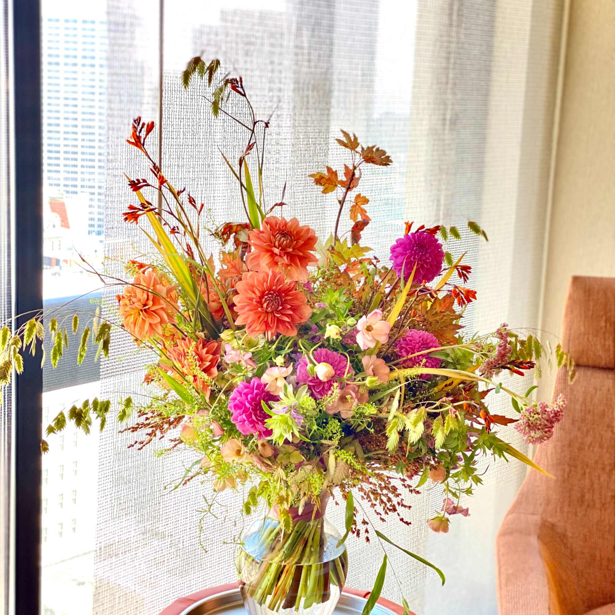 Tall arrangement of orange and pink dahlias and mixed flowers in a clear glass vase by a window.