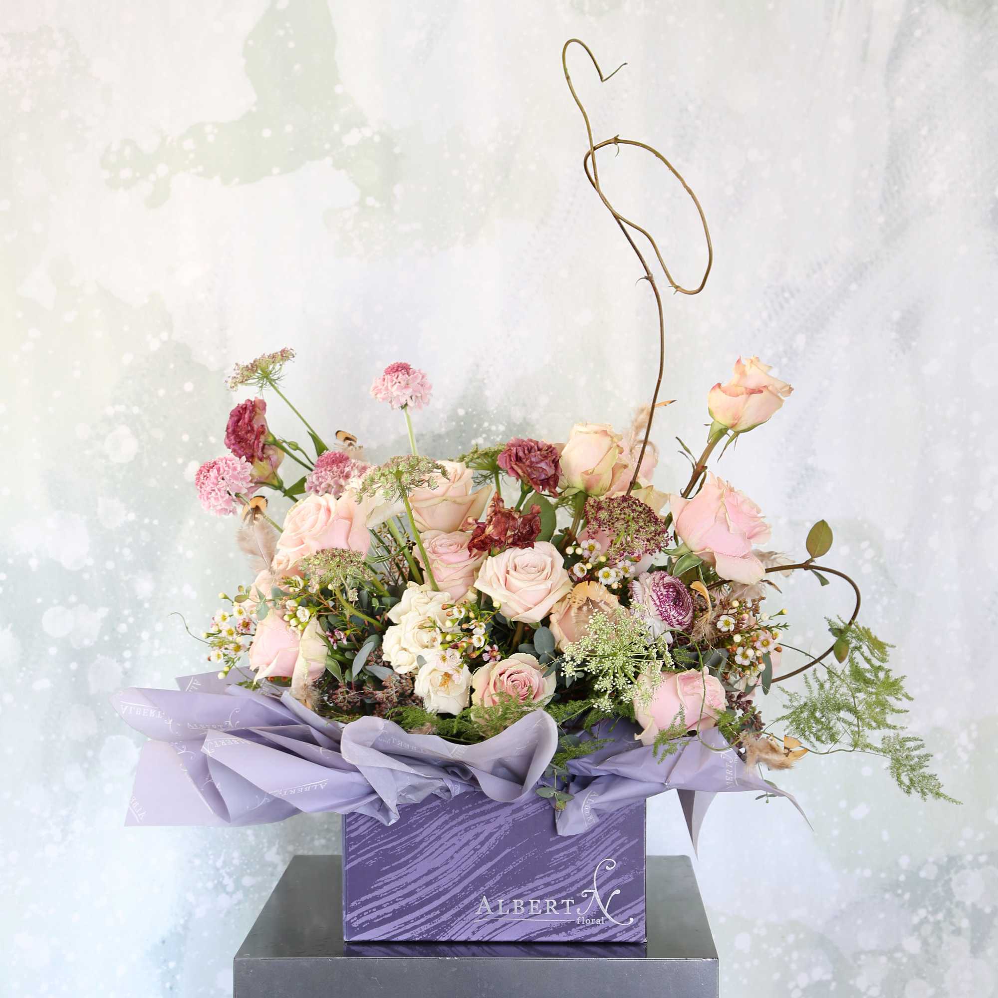 Pastel pink and mauve rose arrangement in a purple box with airy accent blooms and a heart-shaped twig