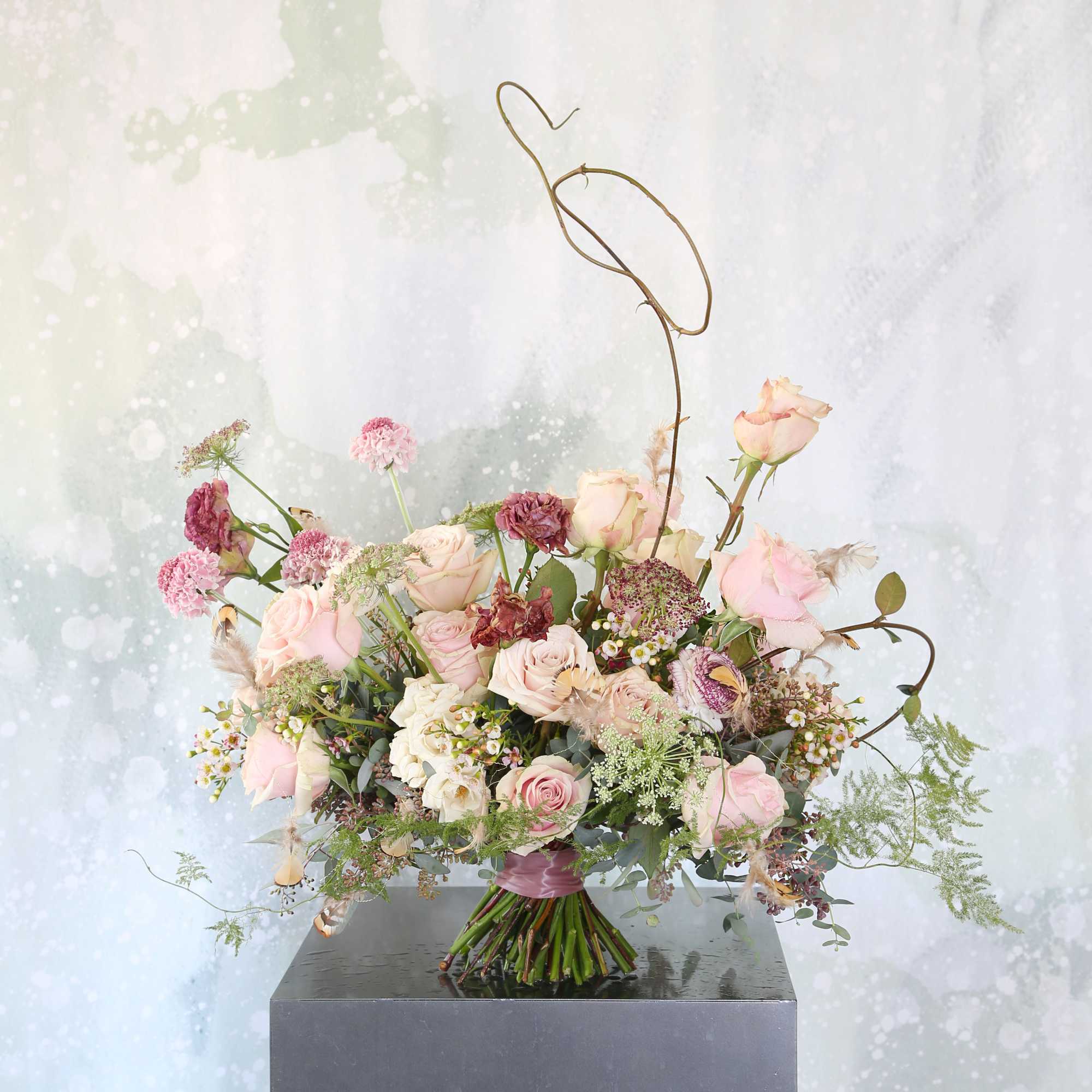 Loose hand-tied bouquet of blush and mauve roses with airy accent flowers and sculptural wire detail.